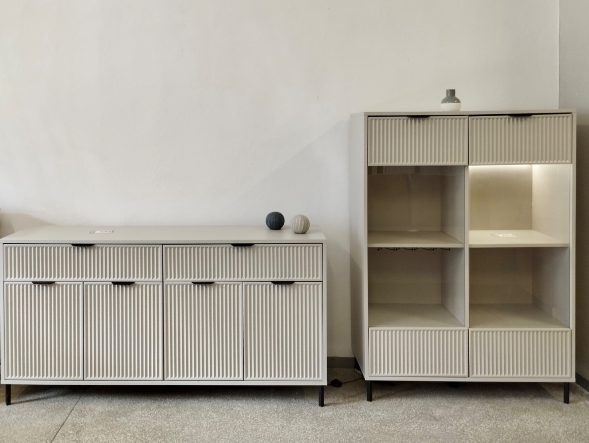 Jasna komoda z ryflowanymi frontami i szafka z podświetlanymi półkami na czarnych nóżkach. Na komodzie dwie dekoracyjne kule. Minimalistyczny design.