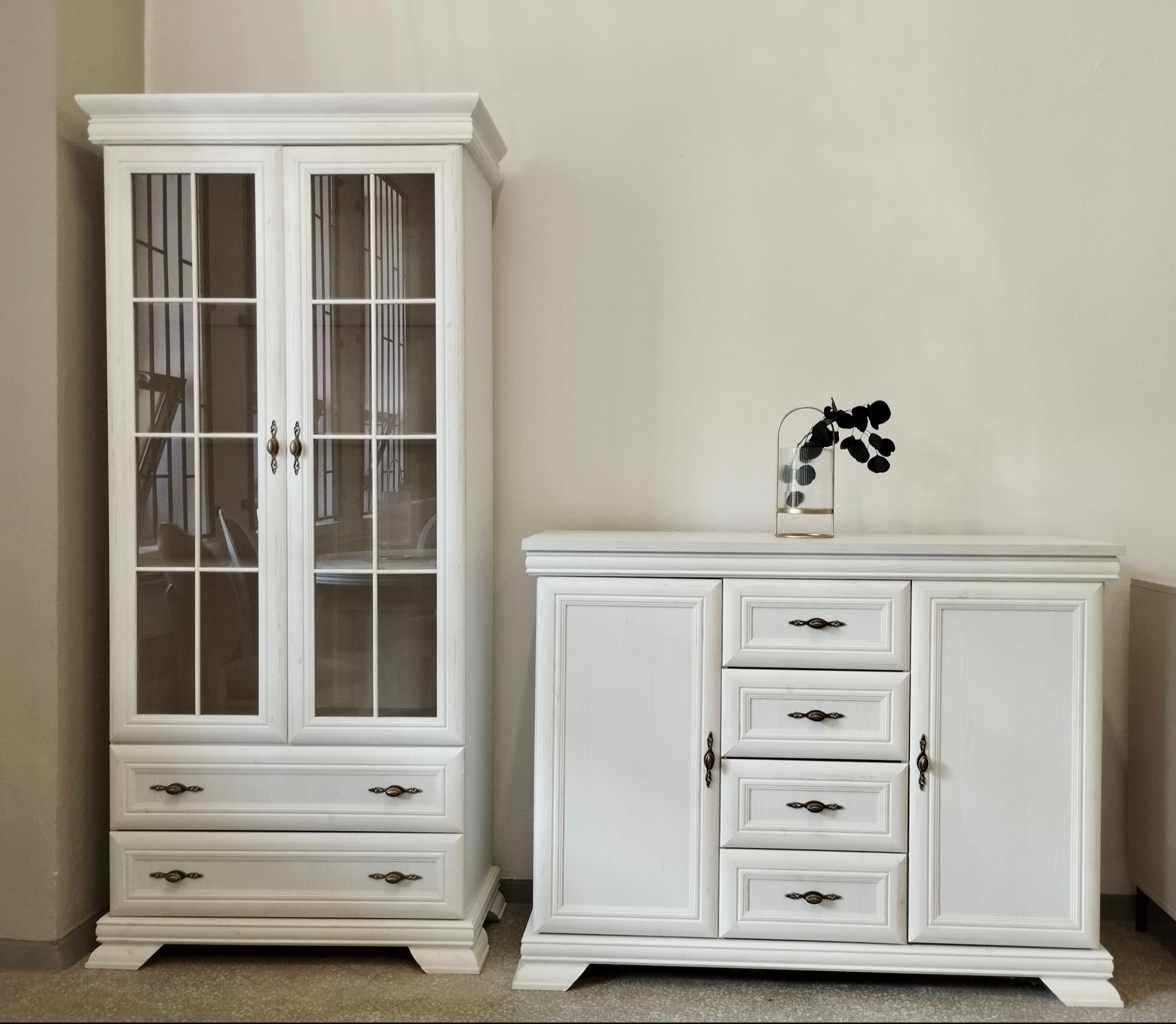 Biała przeszklona witryna z szufladami i komoda z dekoracją. Klasyczny zestaw mebli do salonu lub jadalni, z ciemnymi uchwytami i minimalistycznym wystrojem.