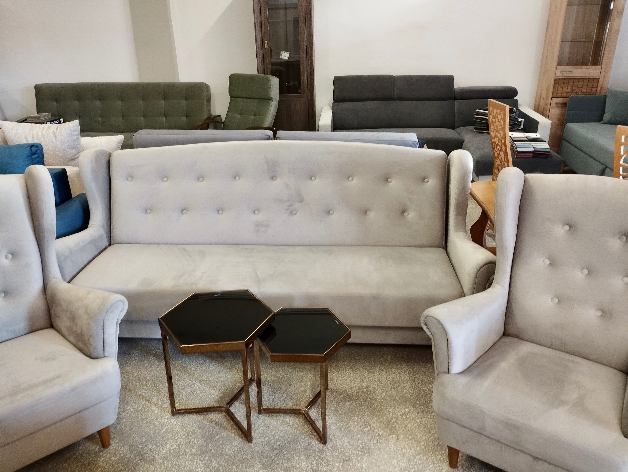 Jasnoszary komplet wypoczynkowy: sofa z guzikami i dwa fotele uszaki, uzupełniony o dwa heksagonalne stoliki kawowe na tle ekspozycji meblowej.