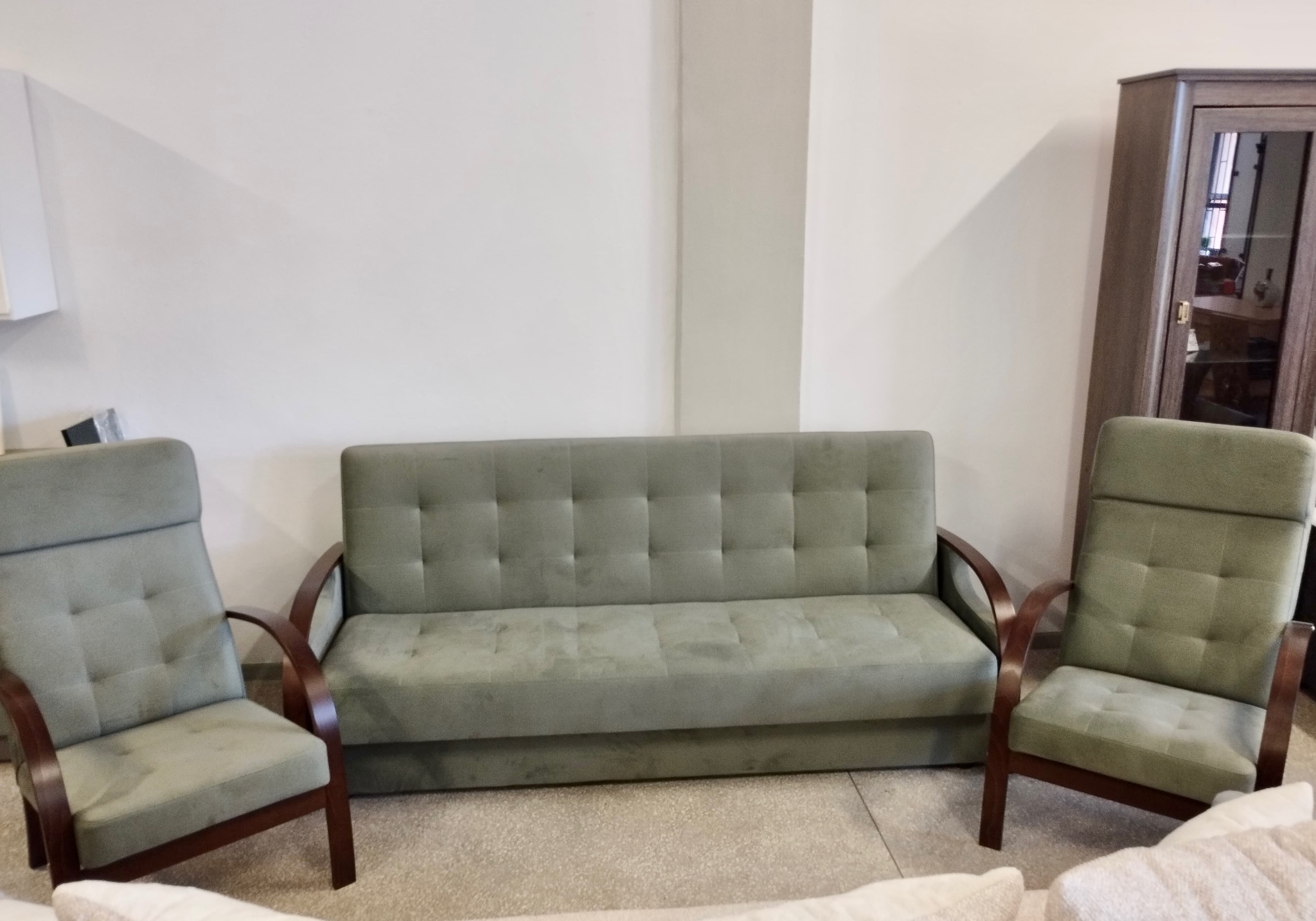 Komplet wypoczynkowy: sofa i dwa fotele w odcieniach zieleni, z drewnianymi elementami, ustawione na tle jasnej ściany i szafy. Widok z bliska, prezentacja mebli.
