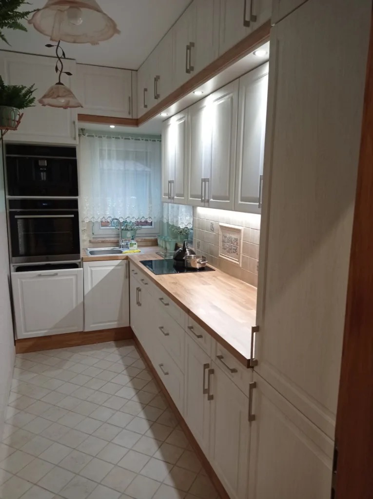 Jasna kuchnia z drewnianym blatem, oświetleniem podszafkowym i zabudowanymi sprzętami. Widok na okno z firanką i dekoracjami.