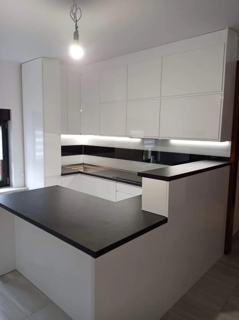 Nowoczesna kuchnia z białymi szafkami i czarnym blatem, oświetlenie LED pod szafkami, minimalistyczny design, widok na wyspę kuchenną.