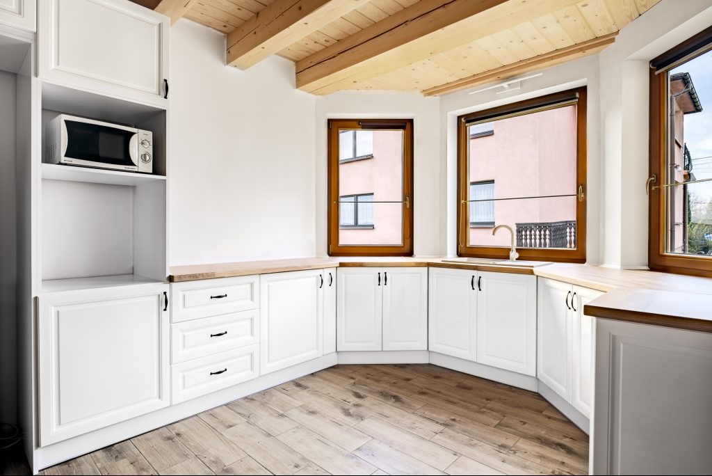 Biała kuchnia z drewnianymi blatami i oknami w brązowych ramach. Podwieszana szafka z mikrofalówką. Drewniany sufit z belkami. Parkiet w jasnym odcieniu drewna.