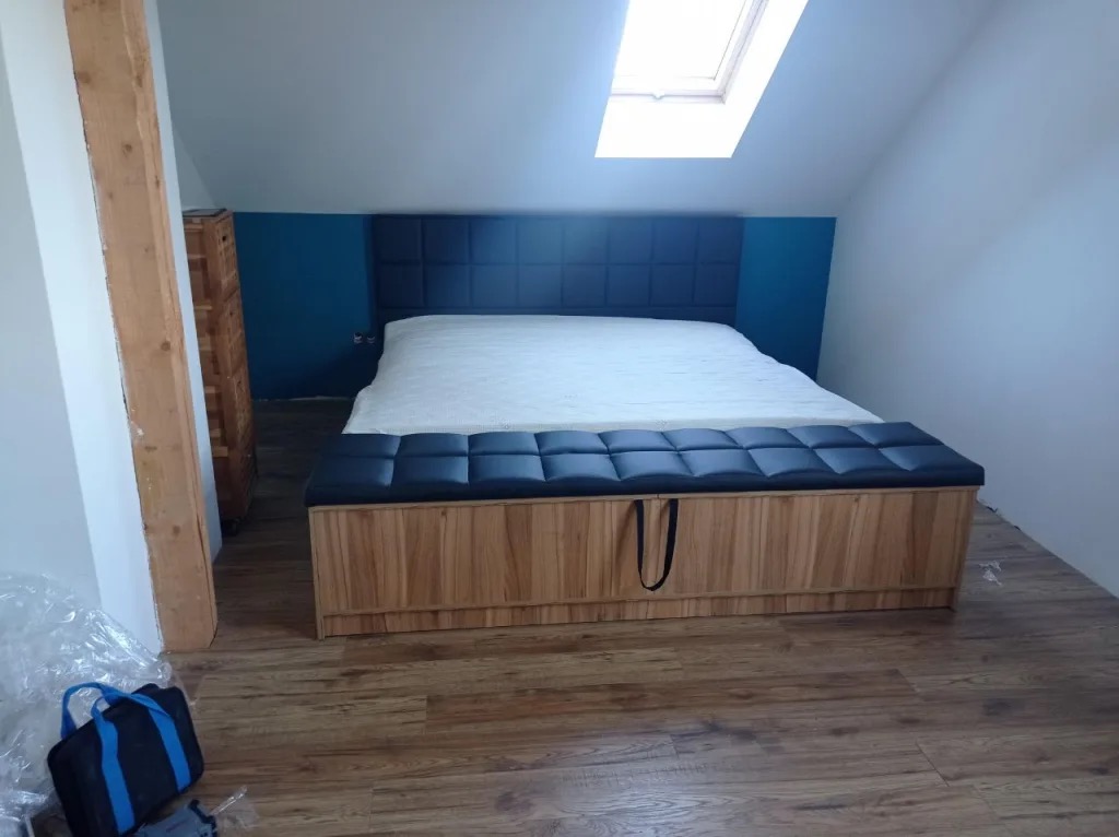 Drewniane łóżko z tapicerowanym wezgłowiem i skrzynią w pokoju na poddaszu z oknem dachowym. Skrzynia obita czarną skórą z uchwytami.
