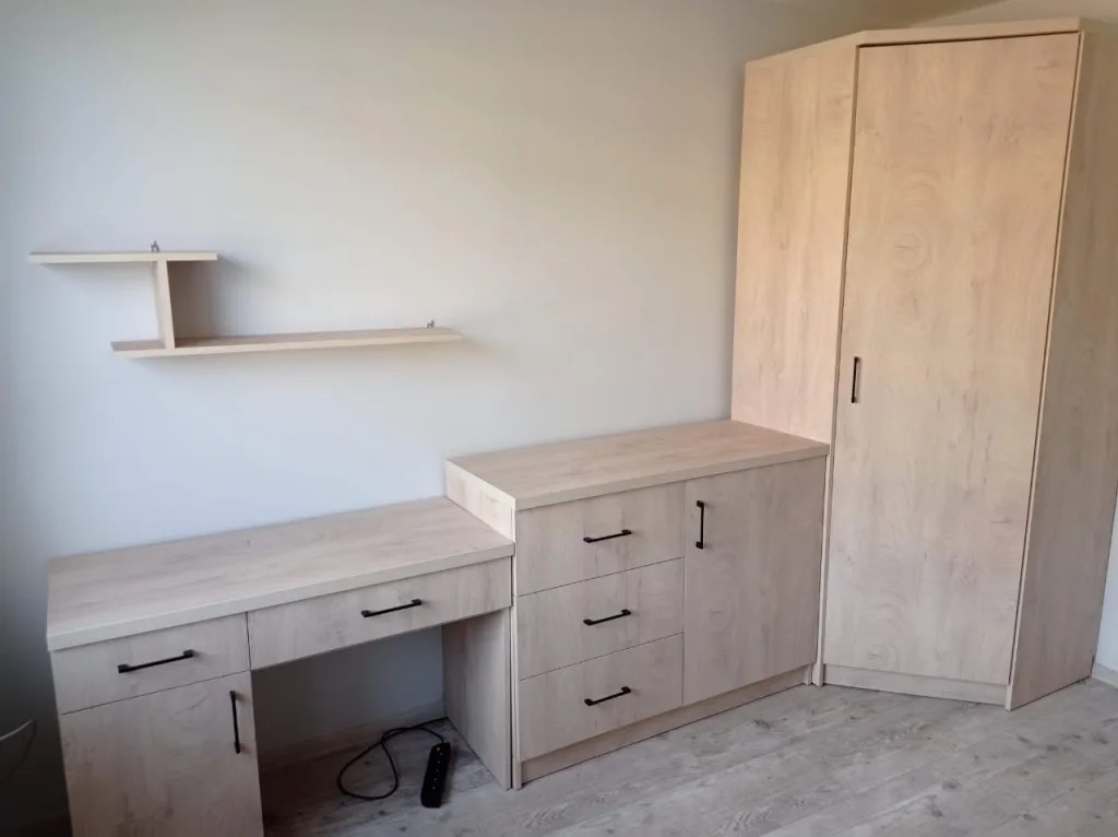 Jasna zabudowa meblowa: szafa narożna, komoda z szufladami i biurko z szafką, uzupełnione półką wiszącą. Czarne uchwyty dodają kontrastu.
