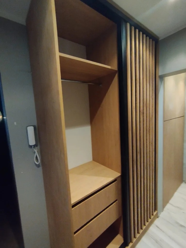 Jasna szafa wnękowa z przesuwnymi drzwiami w pionowe lamele, widoczne półki i szuflady, minimalistyczny design.