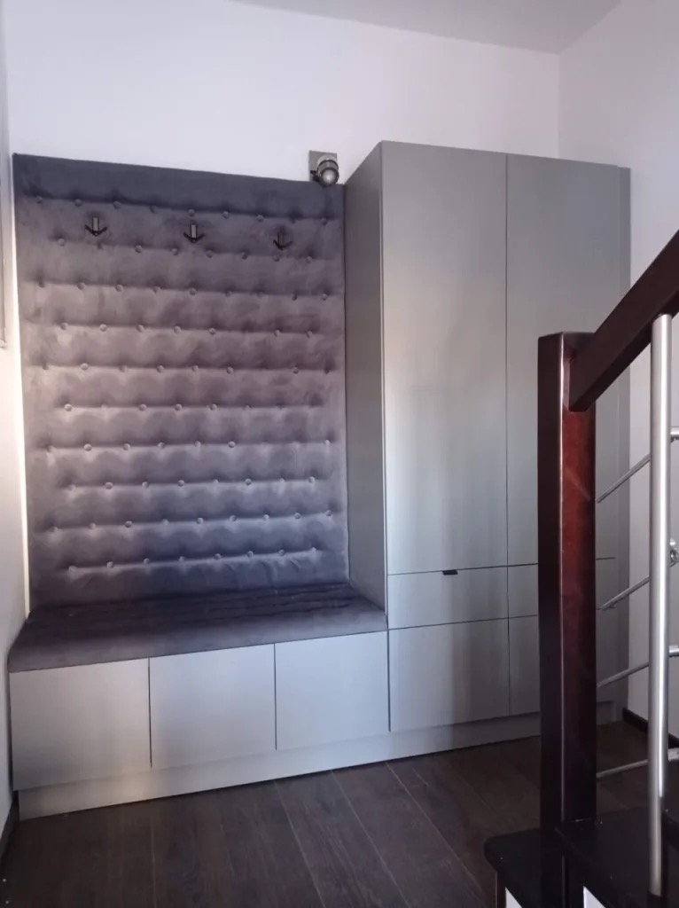 Szara zabudowa wnęki z miękkim siedziskiem i panelami z guzikami oraz wieszakami, obok szafa w tym samym odcieniu, drewniana podłoga i fragment schodów.