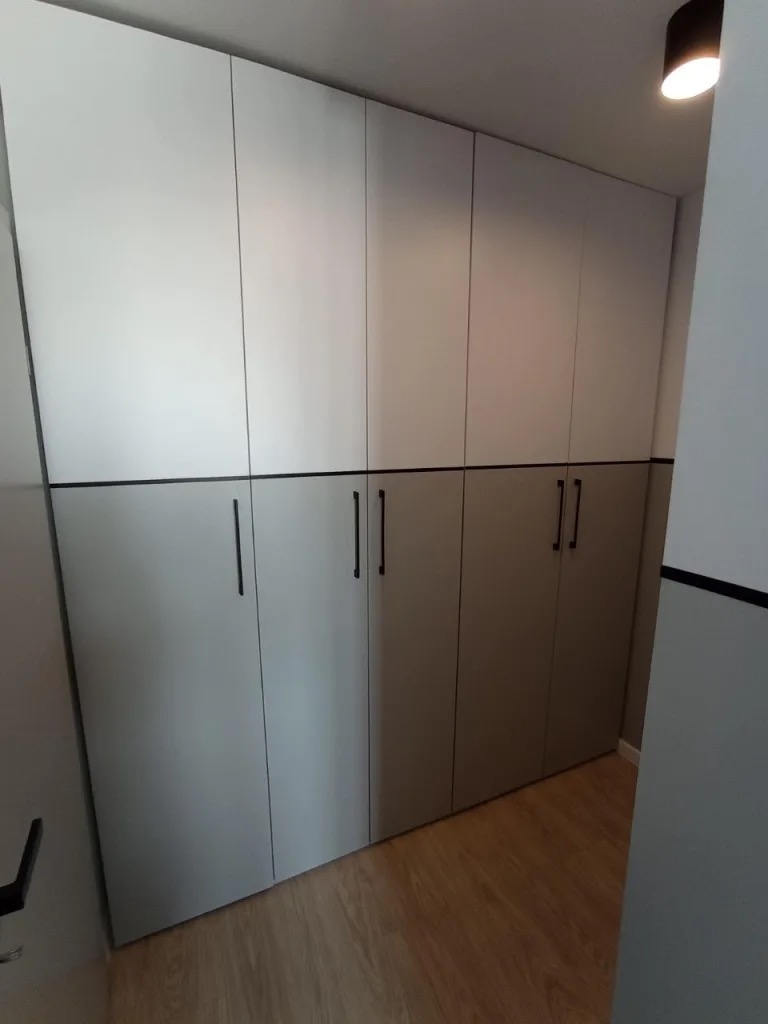 Dwukolorowa szafa wnękowa z długimi, czarnymi uchwytami, z jasną górą i ciemniejszym dołem, na drewnianej podłodze, oświetlona górnym światłem.