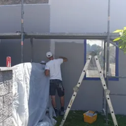 Pracownik w trakcie szpachlowania szarej elewacji domu, stojąc na rusztowaniu. Widoczne narzędzia, okno z niebieską ramą i drzewo. Częściowo zasłonięte folią.