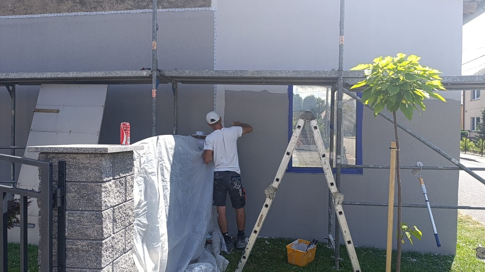 Pracownik w trakcie szpachlowania szarej elewacji domu, stojąc na rusztowaniu. Widoczne narzędzia, okno z niebieską ramą i drzewo. Częściowo zasłonięte folią.