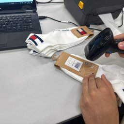 PURELLS INNOVATIVE SPÓŁKA Z OGRANICZONĄ ODPOWIEDZIALNOŚCIĄ - Skanowanie kodu kreskowego na opakowaniu skarpetek Tommy Jeans za pomocą skanera Newland, obok drukarka etykiet i laptop Dell, w procesie pakowania.