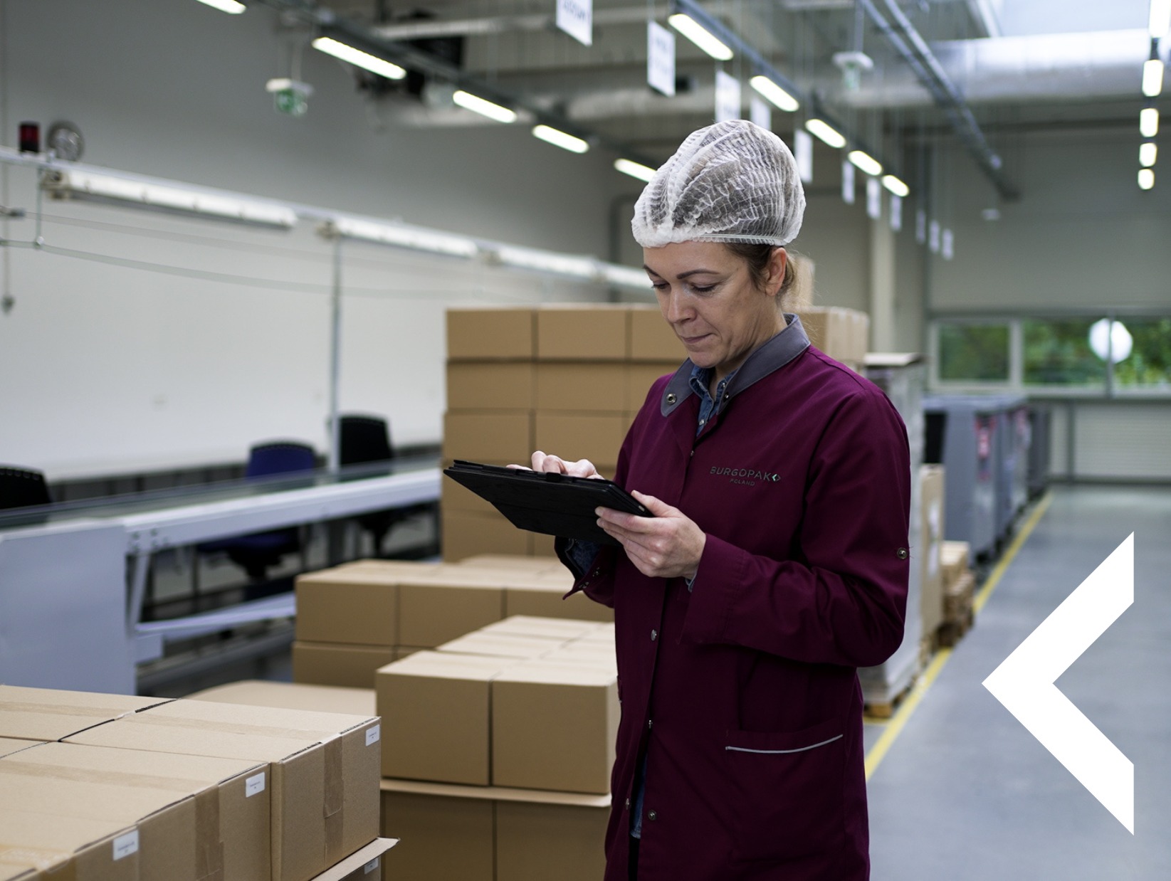 Pracownica w czepku i bordowym uniformie BURGOPAK sprawdza zamówienia na tablecie w hali magazynowej z kartonami i linią produkcyjną w tle.