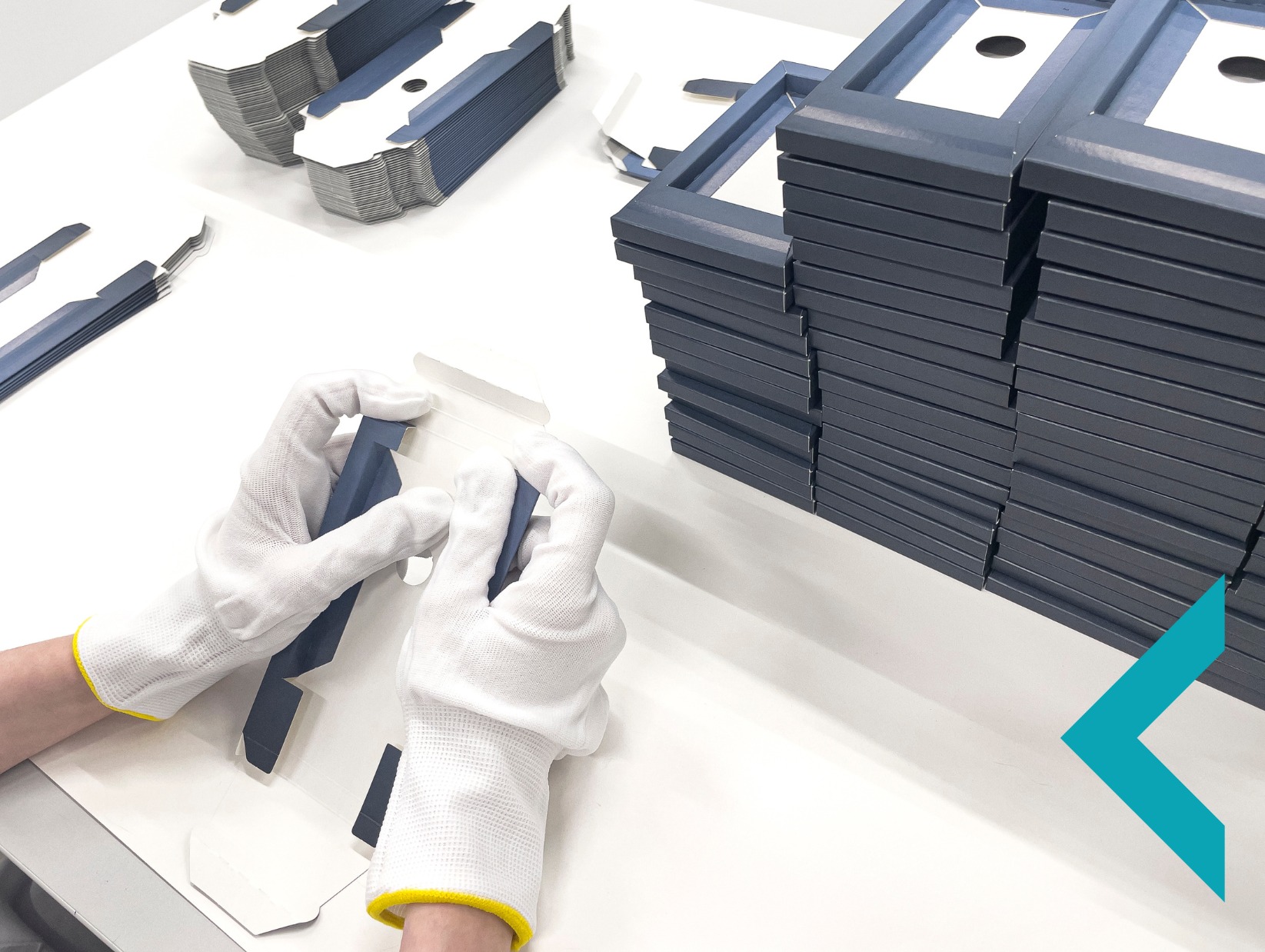 Białe rękawiczki składają pudełko z niebieskiego kartonu. W tle stosy złożonych pudełek i rozłożone elementy. Jasne, sterylne otoczenie.