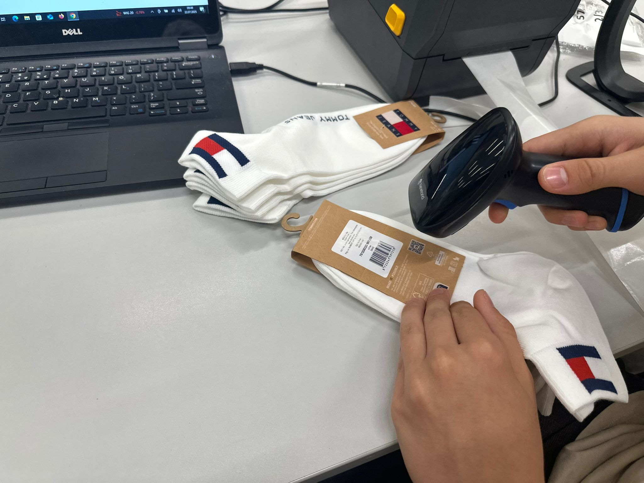 Skanowanie kodu kreskowego na opakowaniu skarpetek Tommy Jeans za pomocą skanera Newland, obok drukarka etykiet i laptop Dell, w procesie pakowania.