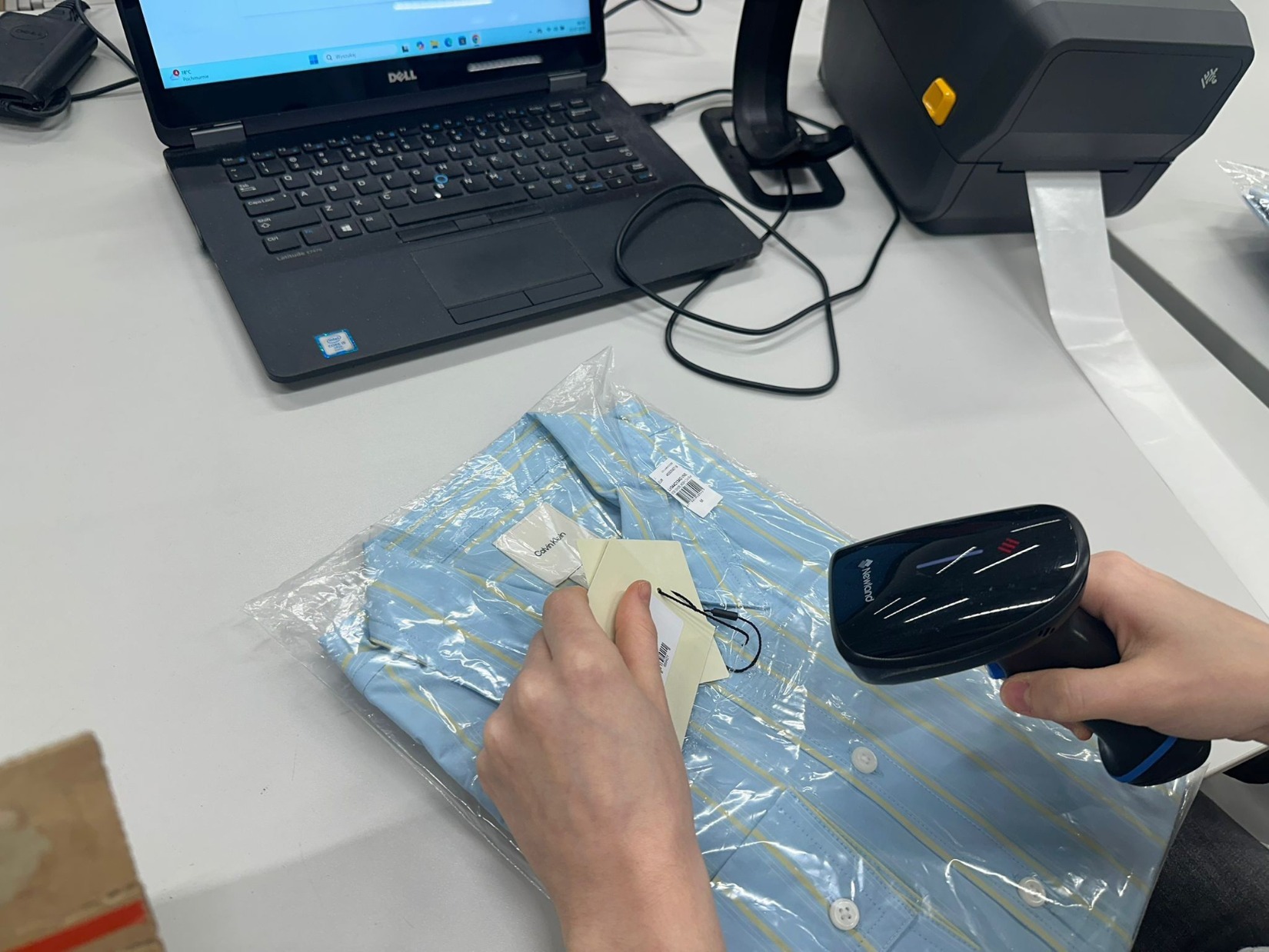 Skanowanie kodu kreskowego koszuli Calvin Klein zapakowanej w folię, obok drukarka etykiet, laptop Dell. Przygotowanie do wysyłki zamówienia.