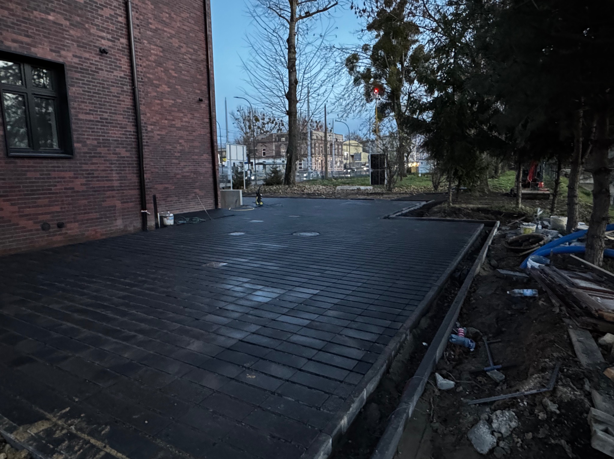 Ułożona, ciemna kostka brukowa przy budynku z czerwonej cegły. Widoczne drzewa w tle i elementy wykończeniowe. Kąt ujęcia z dołu podkreśla regularność wzoru.