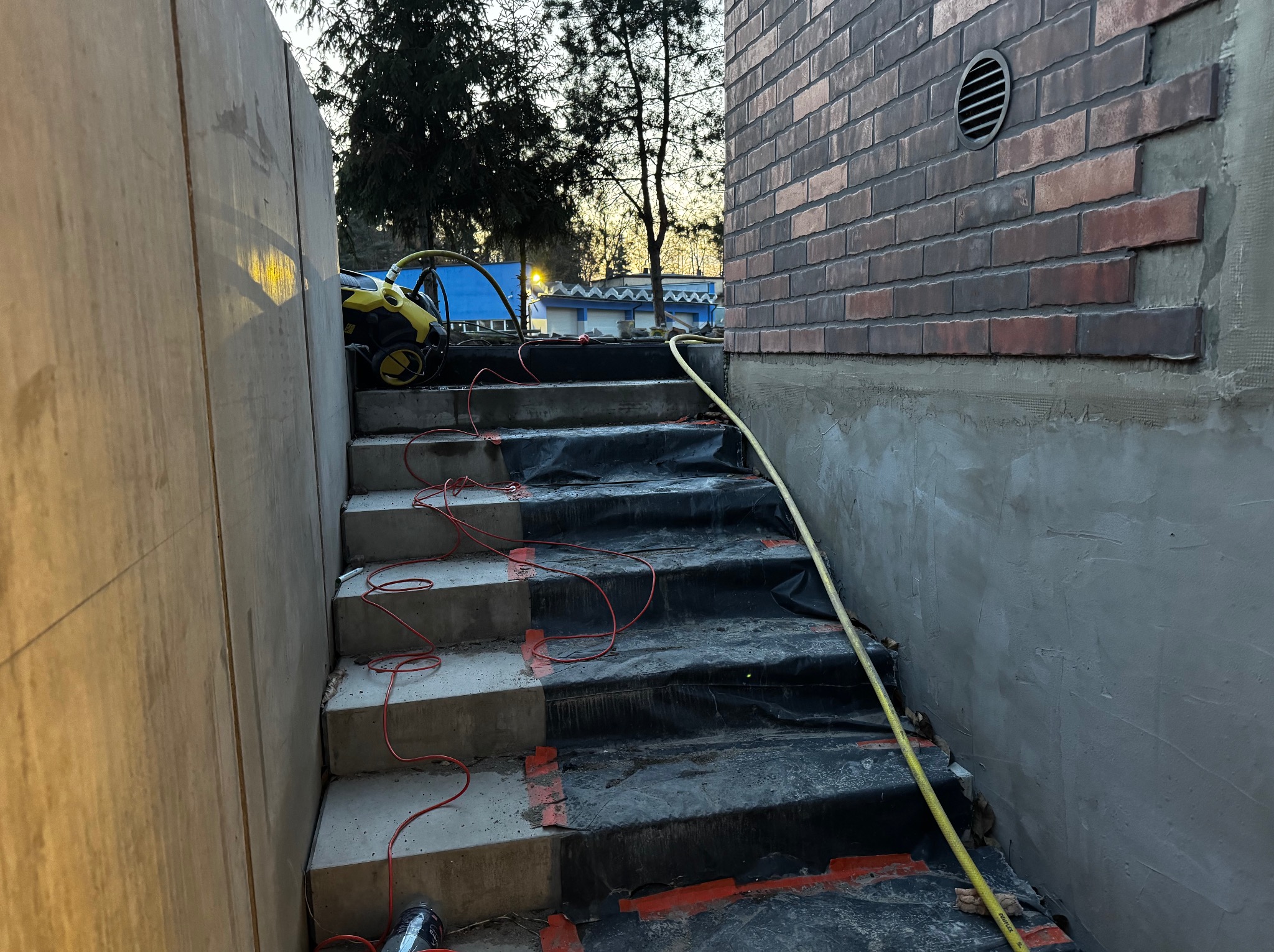 Betonowe schody w trakcie budowy, zabezpieczone folią, z widocznym fragmentem ściany z cegły klinkierowej i żółtym wężem. W tle widoczny sprzęt budowlany.