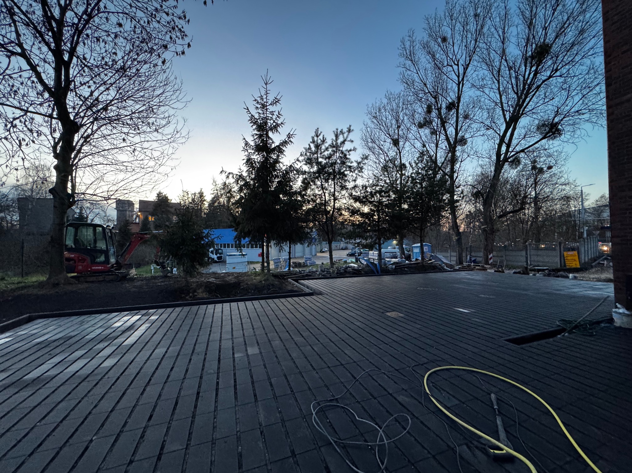 Ciemna, ułożona nawierzchnia z kostki na zewnątrz, z widocznym minikoparką w tle i przewodami na pierwszym planie, oświetlona wieczornym światłem.