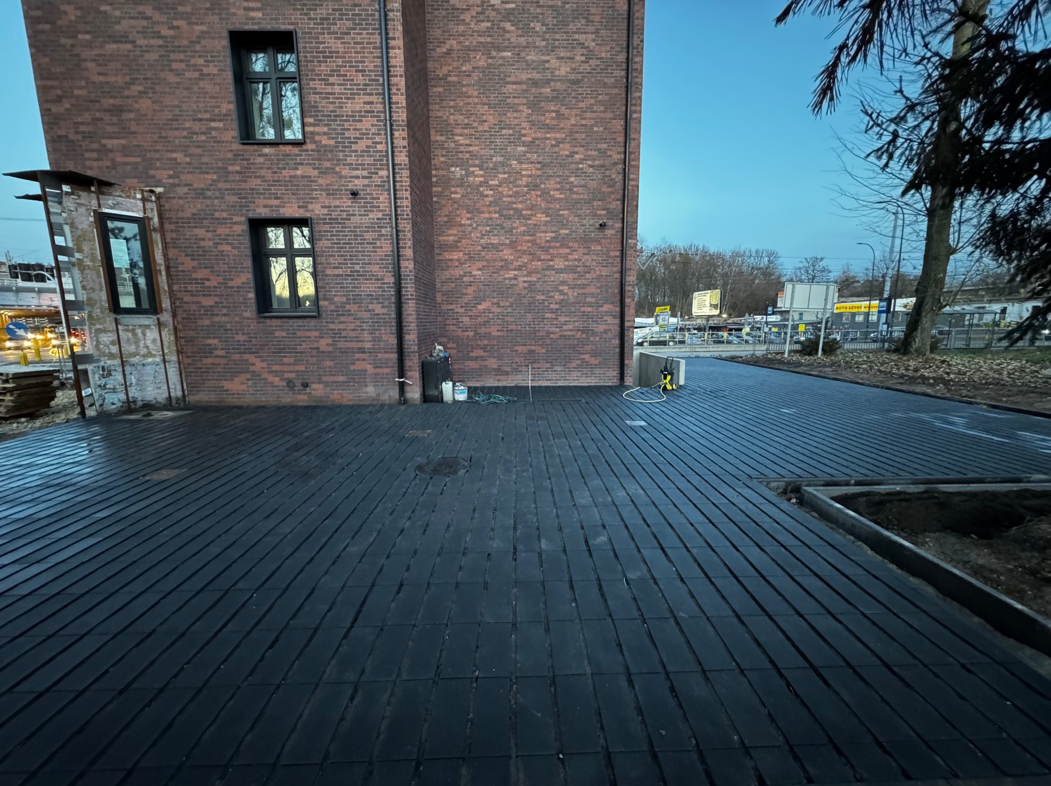 Ułożona, ciemna kostka brukowa przed budynkiem z czerwonej cegły, widoczne narzędzia i resztki materiałów budowlanych. Kąt ujęcia z dołu podkreśla perspektywę.