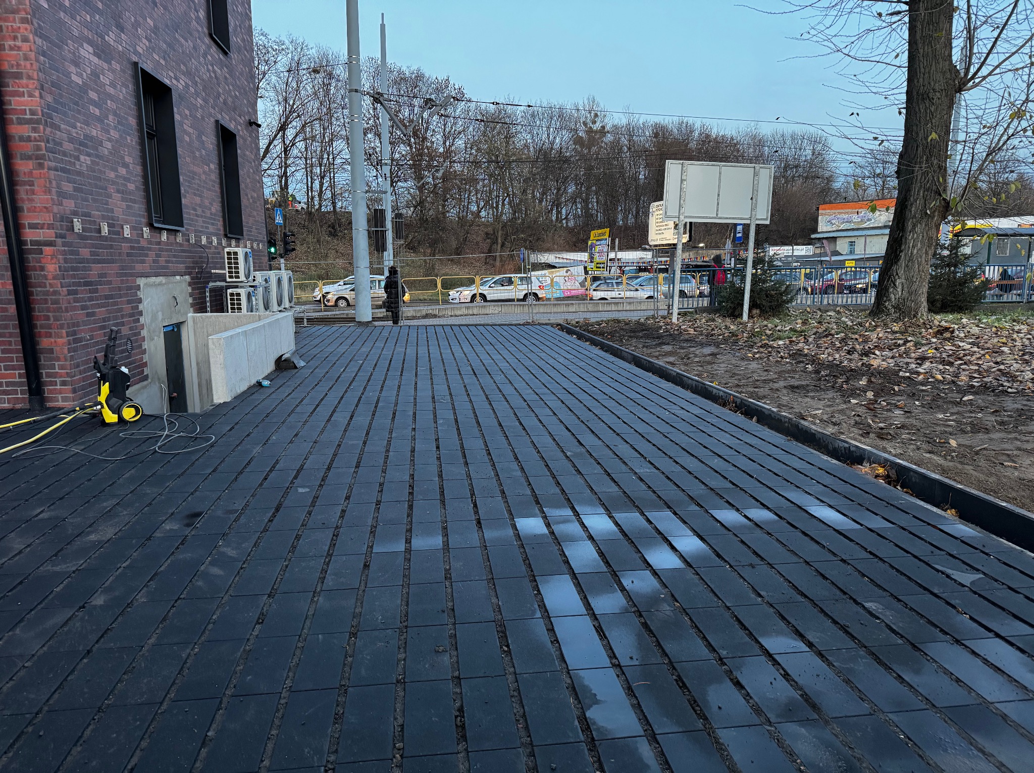 Ułożona, ciemna kostka brukowa wzdłuż budynku z cegły. Widoczny fragment chodnika, ulica w tle i myjka ciśnieniowa. Kąt ujęcia z dołu podkreśla perspektywę.