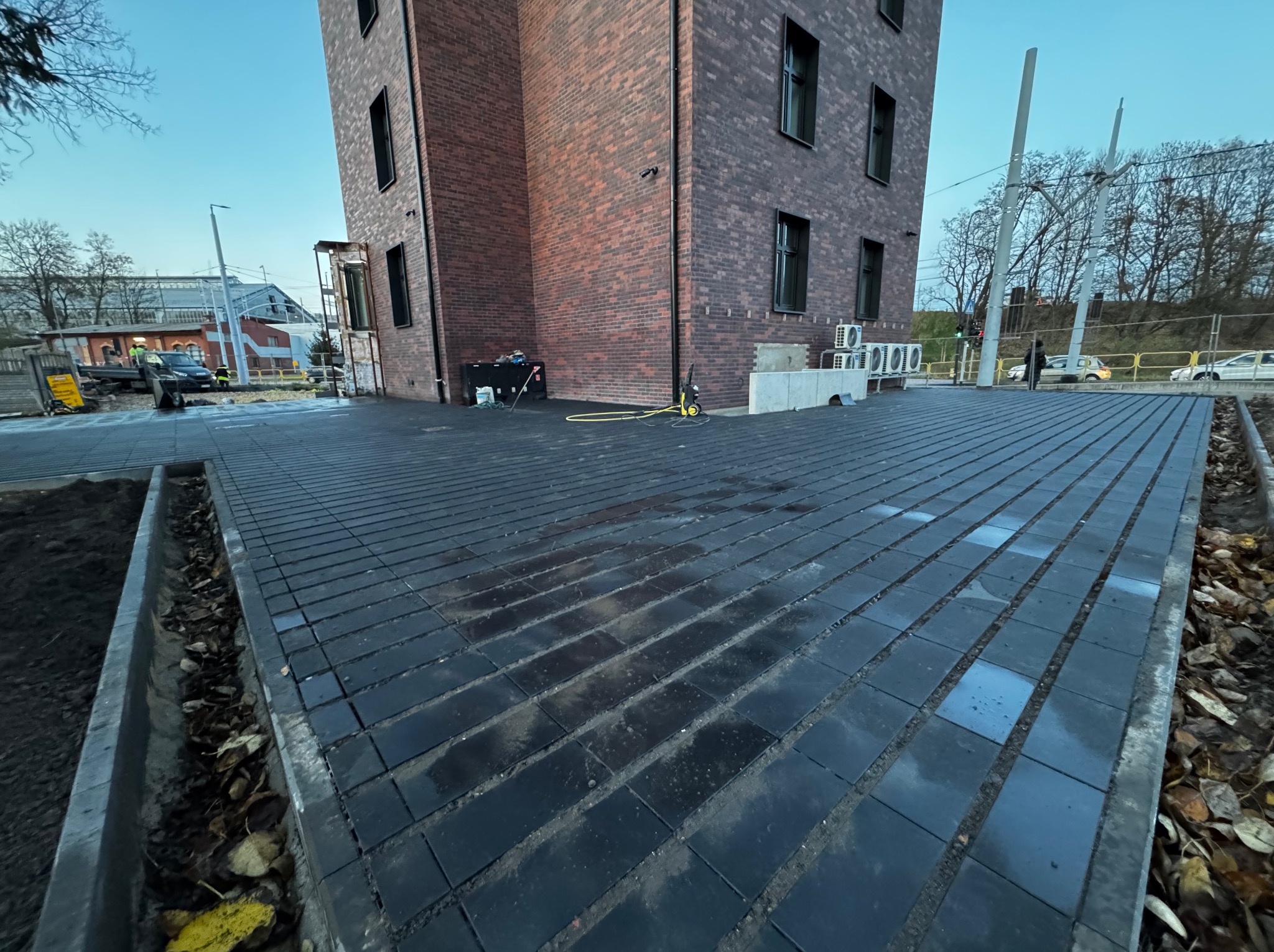 Ułożona, ciemna kostka brukowa przed budynkiem z czerwonej cegły. Widoczne ślady wody na kostce, krawężniki i przygotowane podłoże pod nasadzenia.