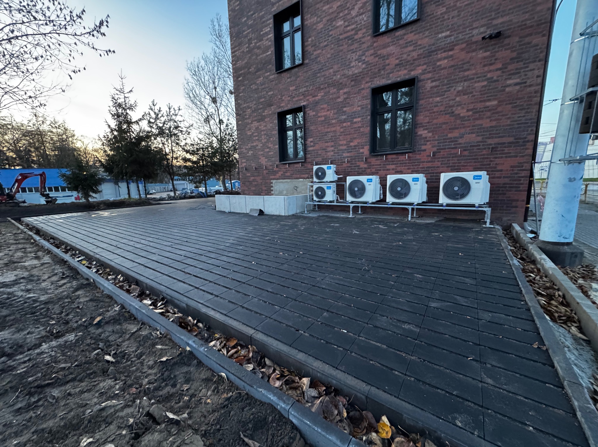 Ułożona, ciemna kostka brukowa przed budynkiem z czerwonej cegły. Widoczne klimatyzatory na zewnątrz i przygotowany teren pod dalsze prace.