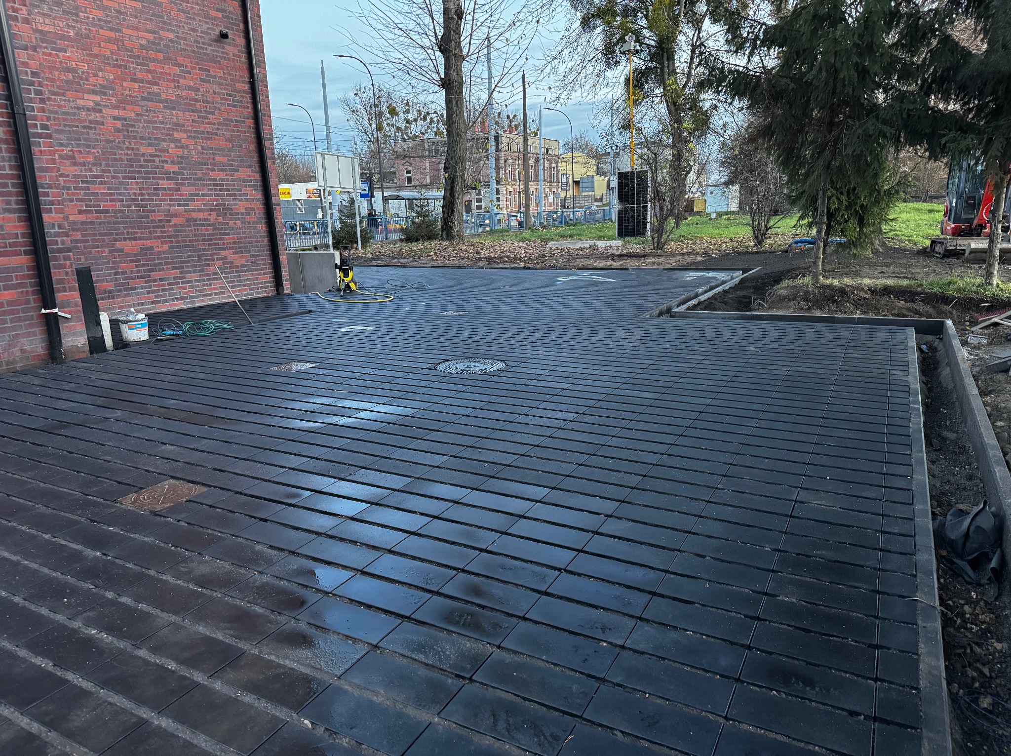 Ułożona, czarna kostka brukowa na posesji, widoczny fragment budynku z czerwonej cegły, maszyna czyszcząca i przygotowany teren pod dalsze prace.