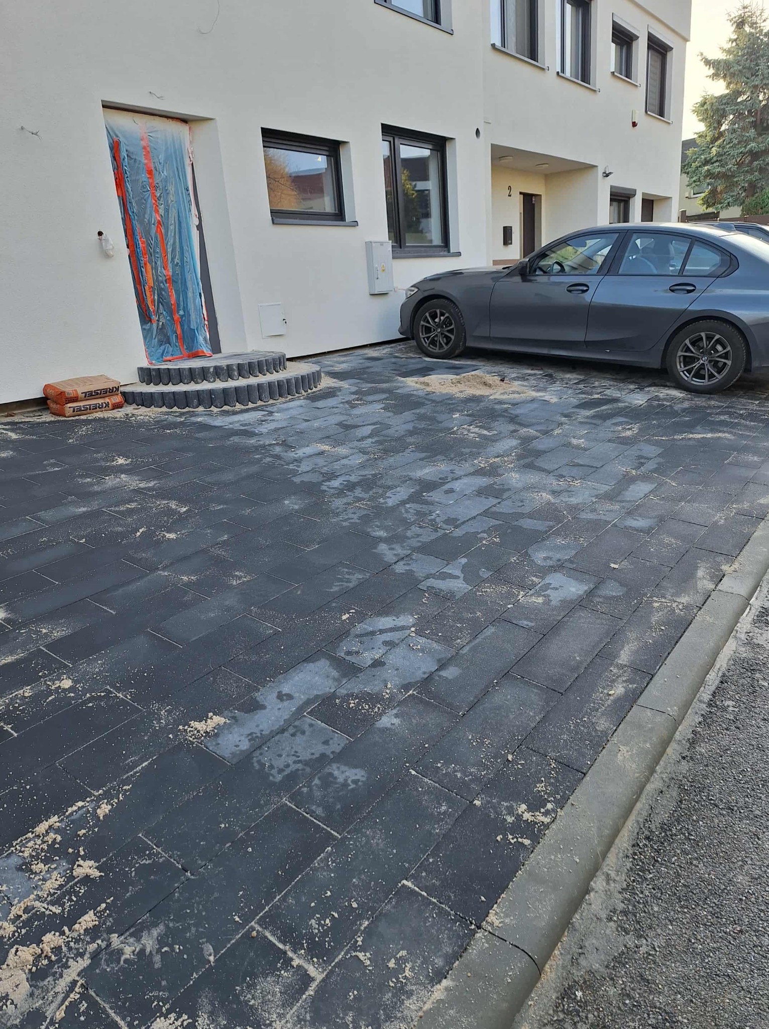 Ułożona, grafitowa kostka brukowa na podjeździe w Chorzowie. Widoczne obrzeża, schody wejściowe i zaparkowany samochód. Częściowo zabezpieczone wejście do domu.