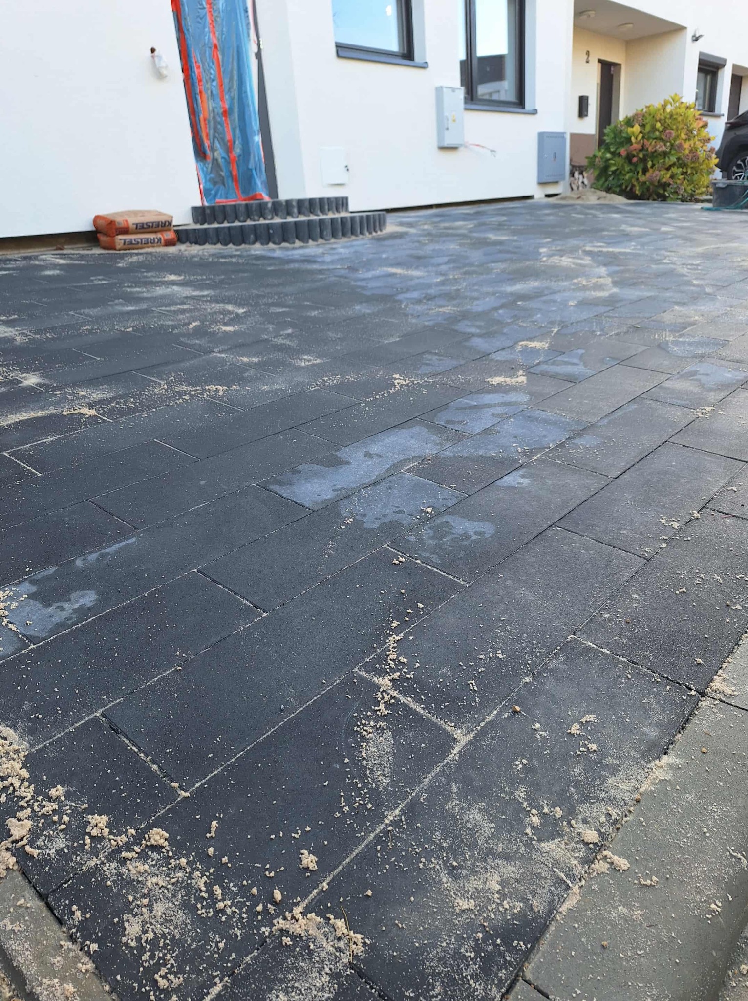 Ułożona, grafitowa kostka brukowa na podjeździe domu jednorodzinnego. Widoczne worki z zaprawą i schody wejściowe. Całość posypana piaskiem.
