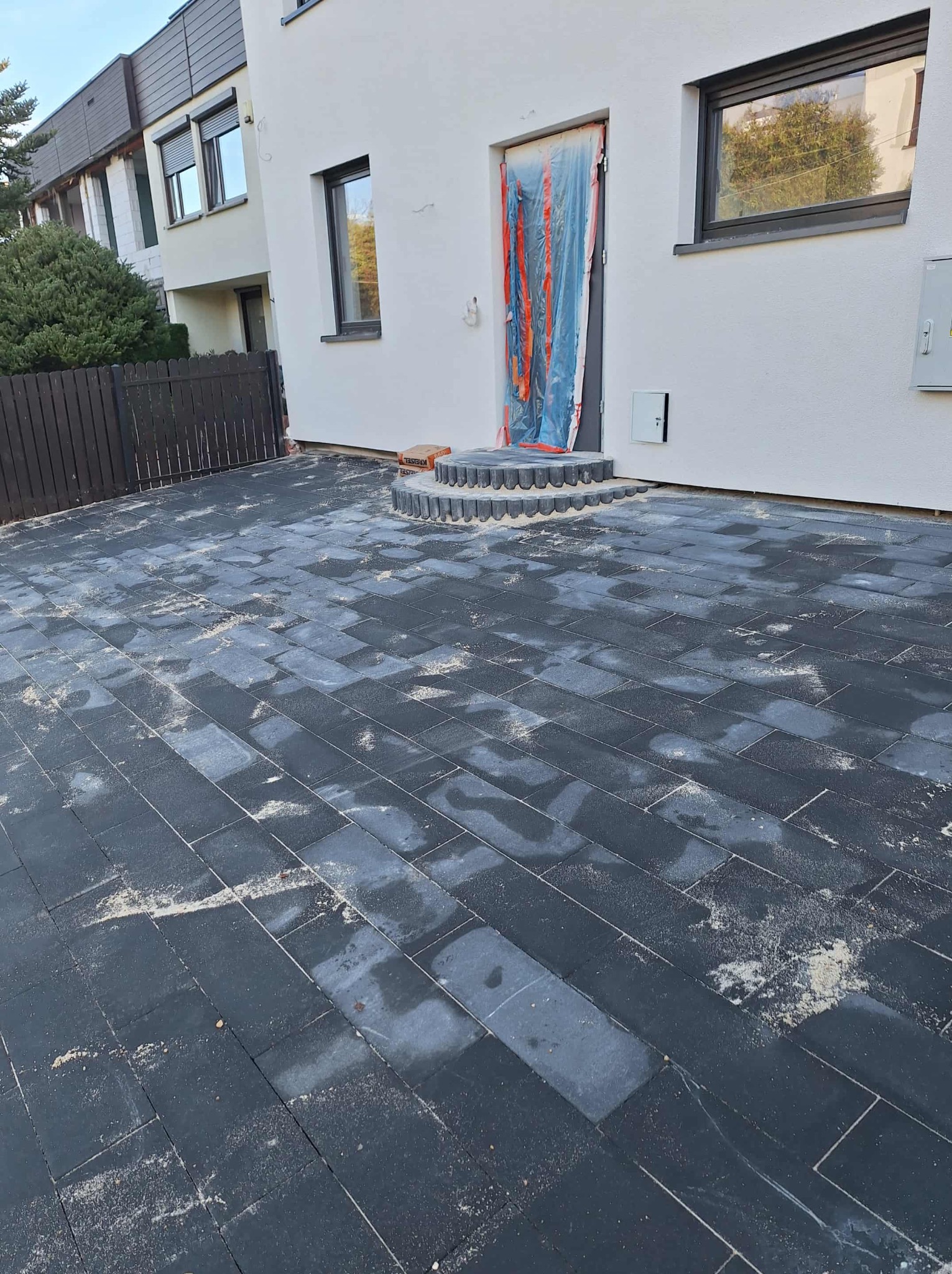 Ułożona, ciemna kostka brukowa przed budynkiem z zabezpieczonym wejściem. Widoczne schody z obrzeżami i piasek na kostce. Ujęcie z dołu, podkreślające wzór.