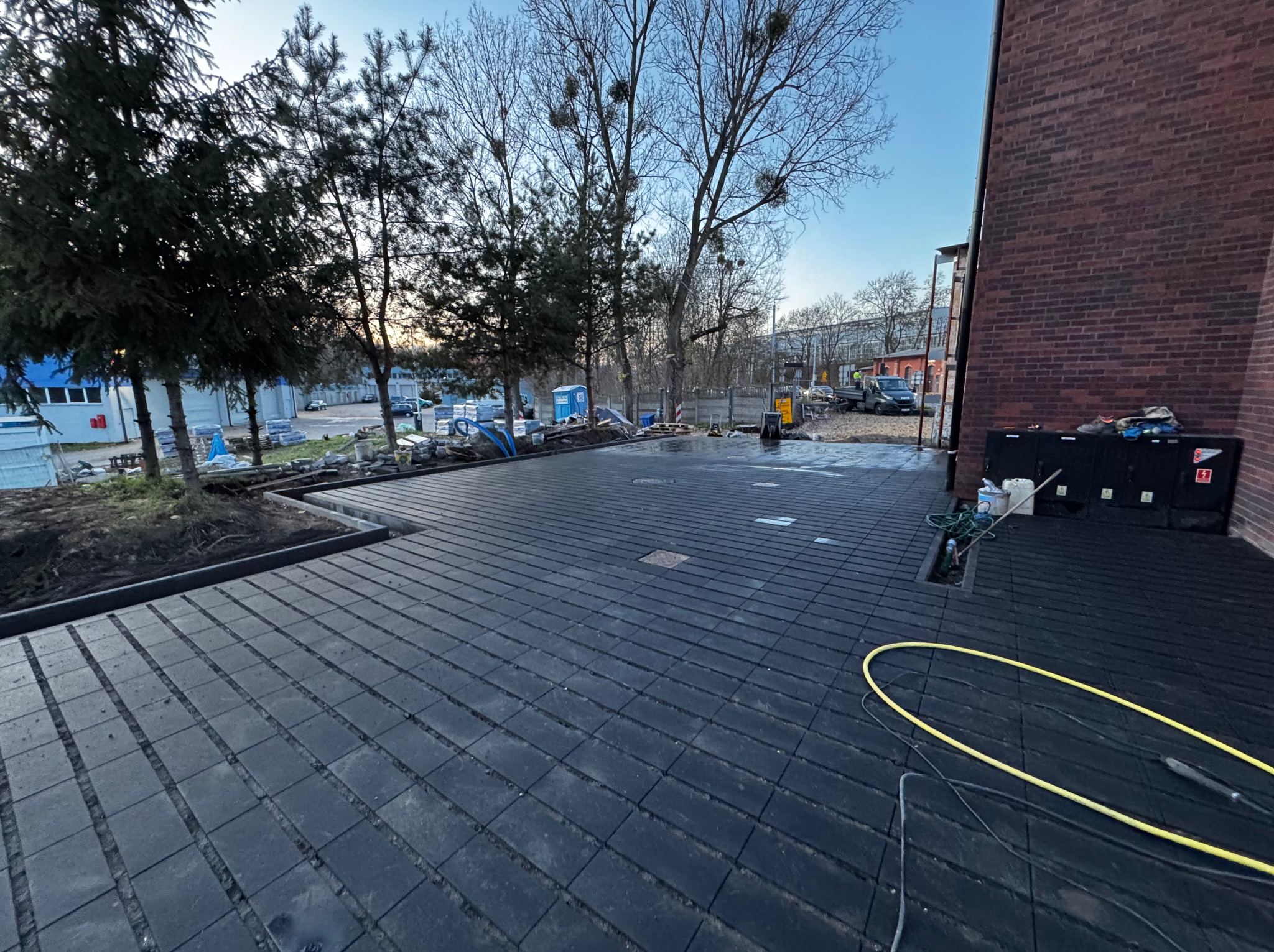 Ułożona, ciemna kostka brukowa na posesji przy budynku z czerwonej cegły. Widoczne drzewa i sprzęt budowlany w tle. Żółty wąż leży na kostce.