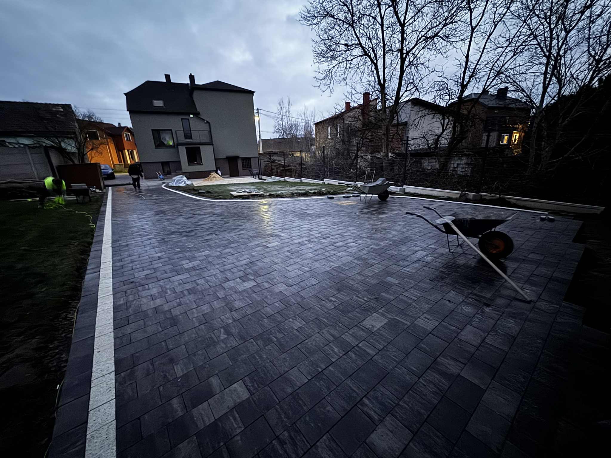 Ukończona nawierzchnia z ciemnej kostki brukowej ułożonej w jodełkę, z białym obrzeżem. W tle budynek mieszkalny i częściowo przygotowany teren pod trawnik.