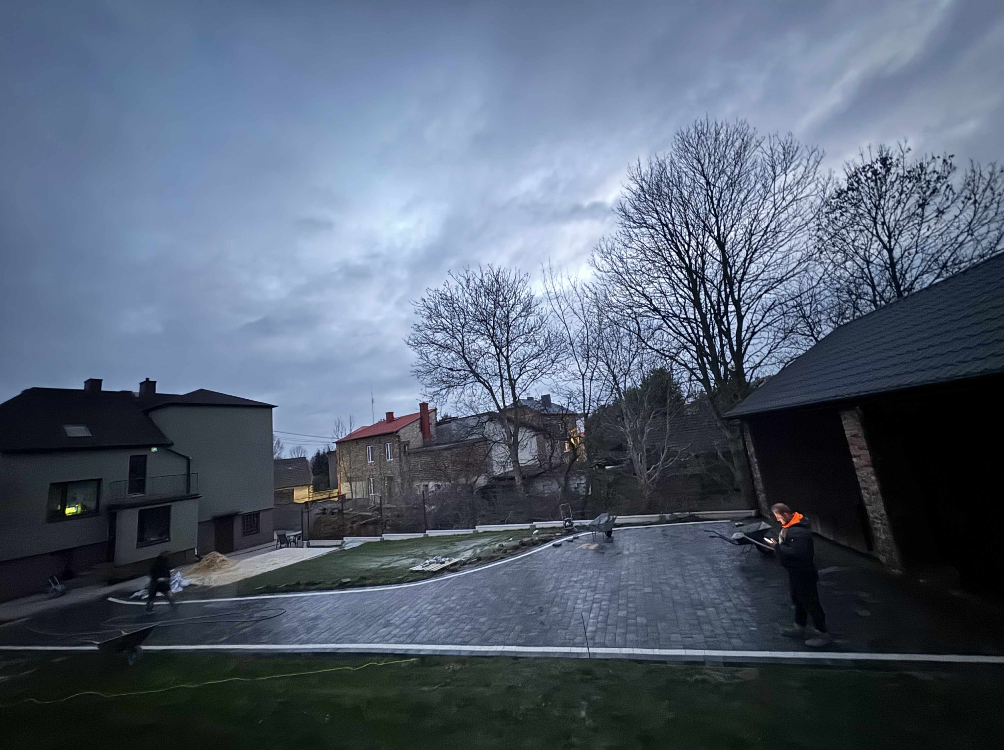 Układanie ciemnej kostki brukowej na posesji, widoczne budynki w tle i pracownicy. Krawędzie kostki wykończone jasnym obramowaniem. Pochmurne niebo.