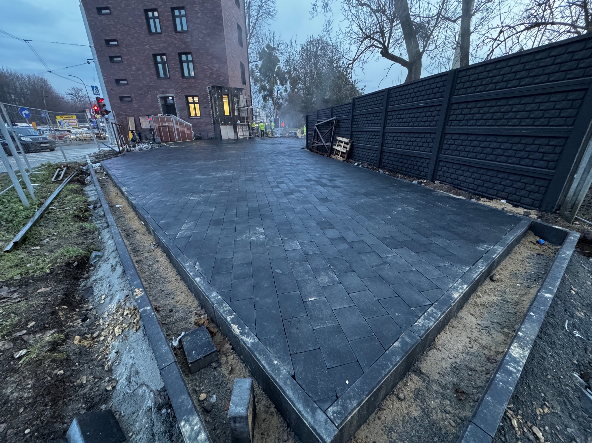 Ułożona, ciemnoszara kostka brukowa na posesji, obrzeża wykończone. W tle budynek i fragment ogrodzenia. Widoczne ślady prac ziemnych.