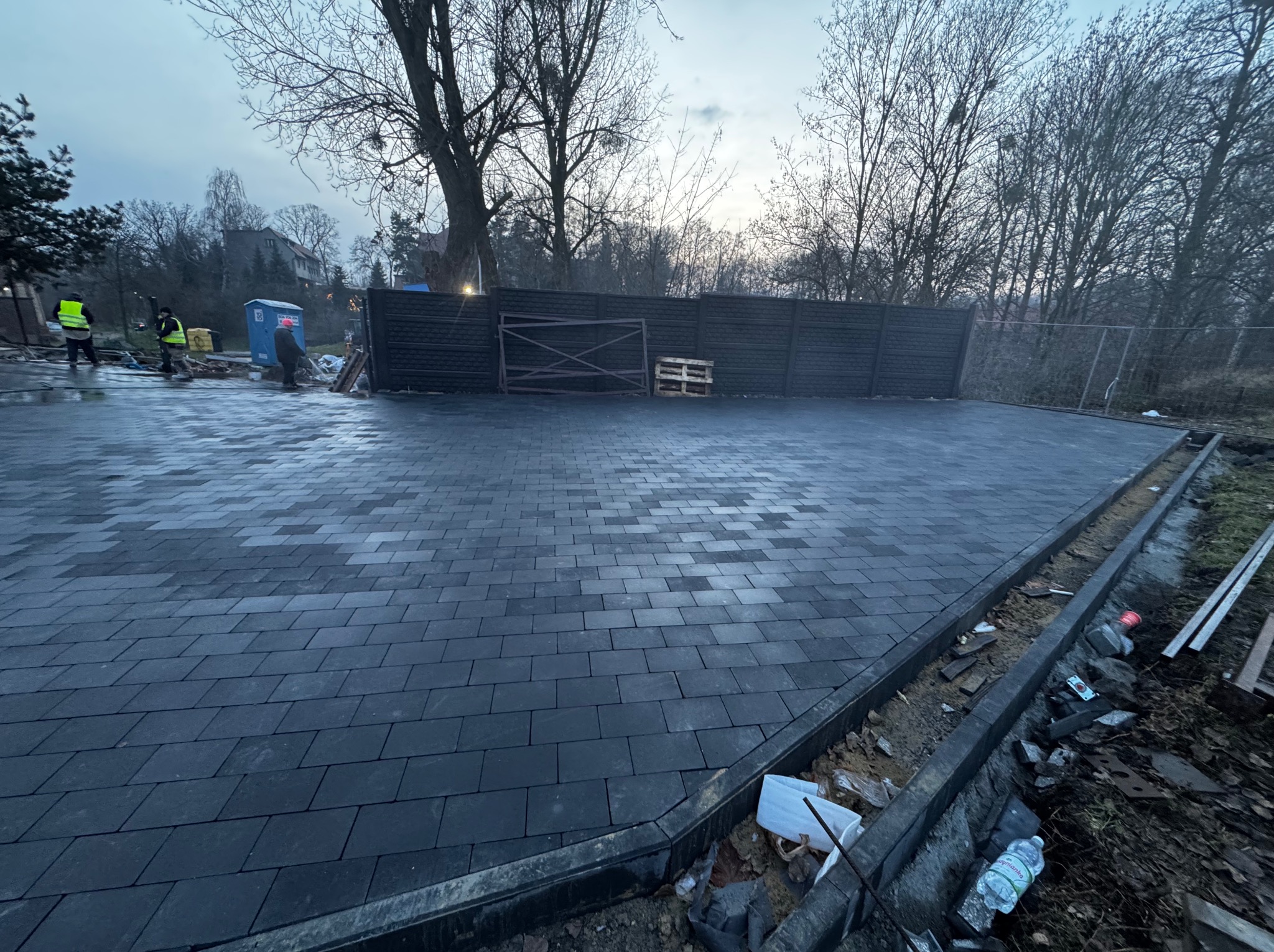 Ułożona, grafitowa kostka brukowa na posesji, widoczny fragment ogrodzenia i paleta. W tle pracownicy w kamizelkach odblaskowych i przenośna toaleta.