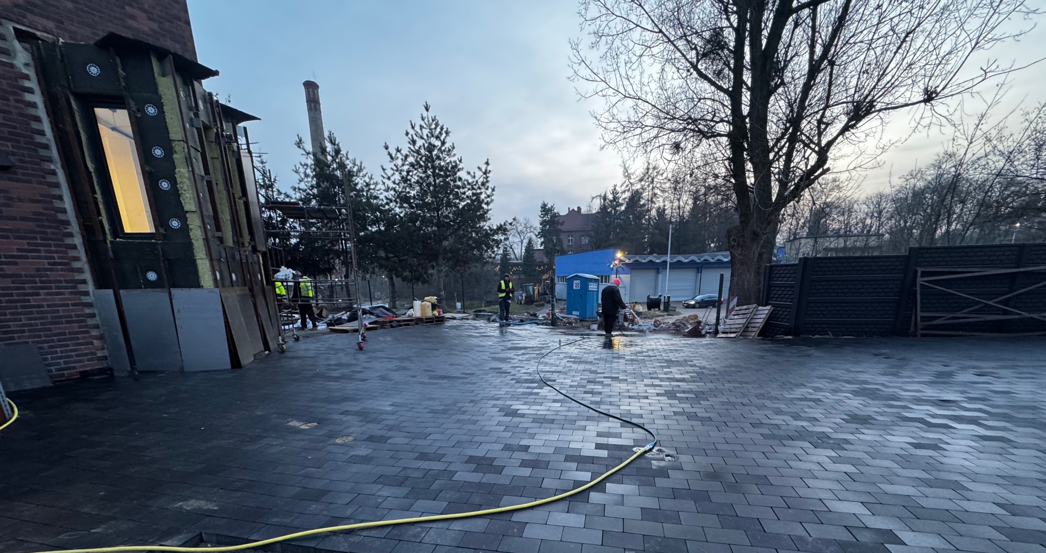 Ułożona, ciemnoszara kostka brukowa na posesji z budynkiem w trakcie remontu. Widoczni pracownicy w odblaskowych kamizelkach i elementy rusztowania.