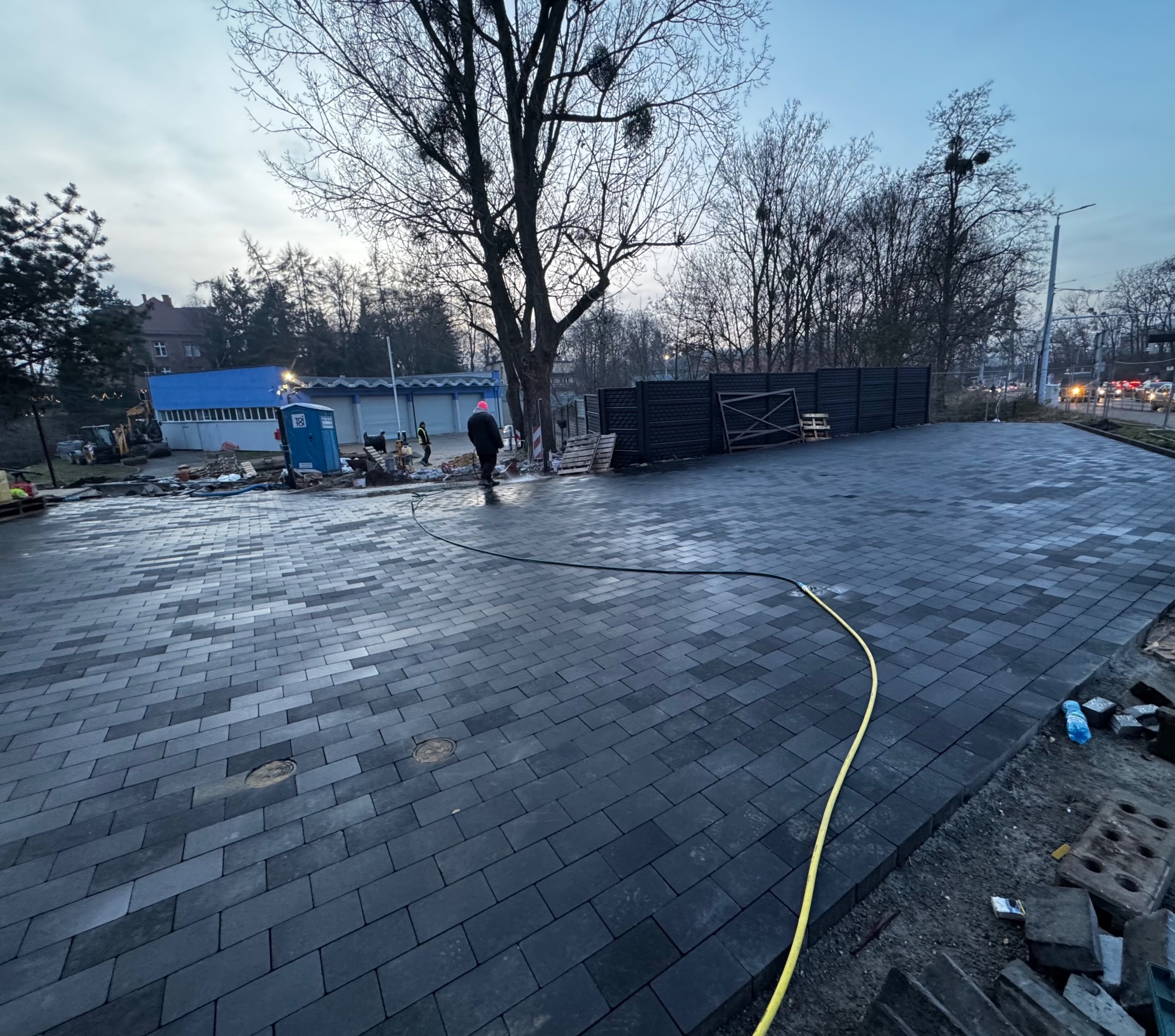 Ułożona, ciemnoszara kostka brukowa na dużej powierzchni, z widocznym fragmentem czarnego ogrodzenia panelowego i pracownikami w tle. W tle budynek i drzewa.
