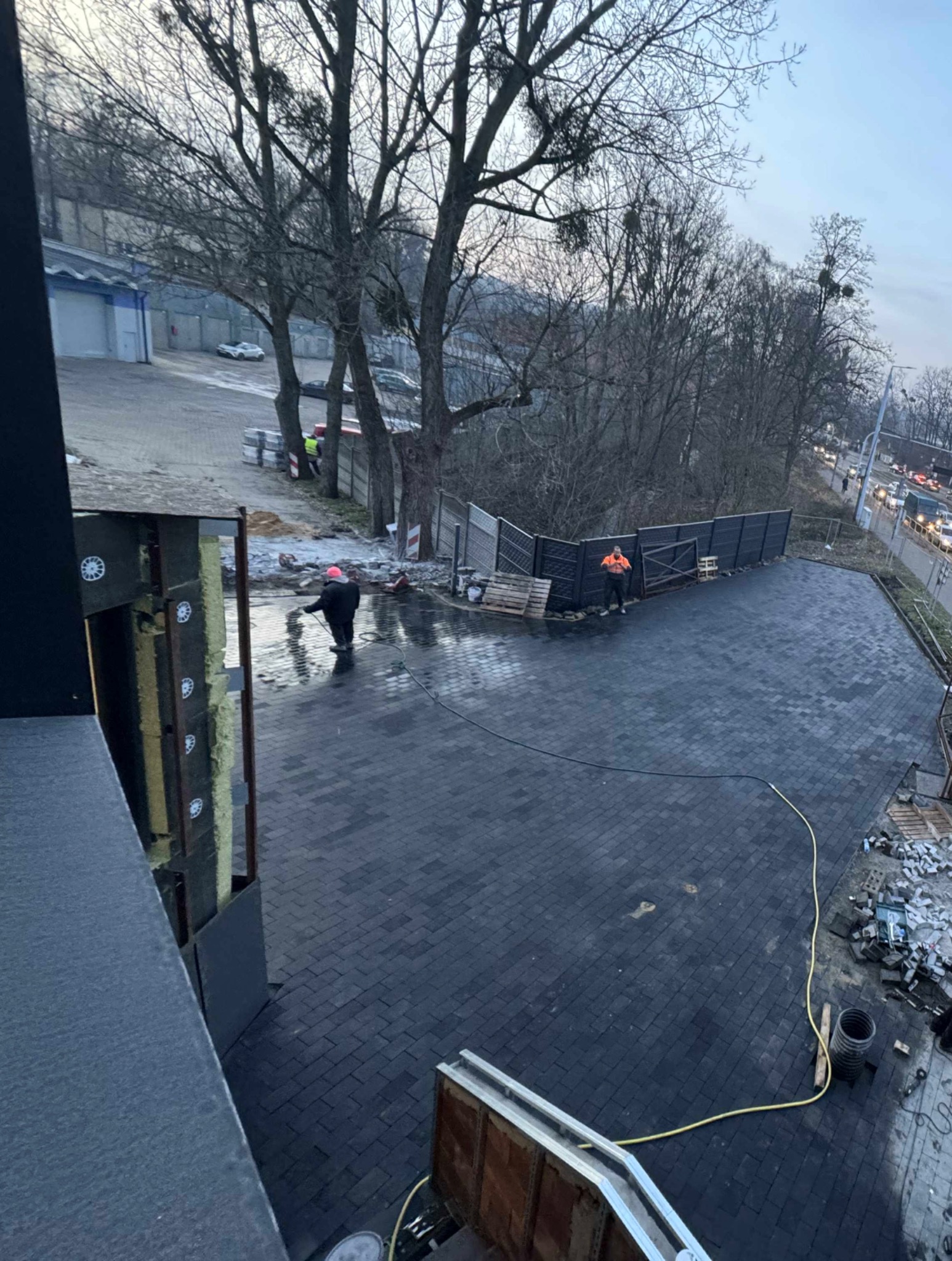 Ciemna kostka brukowa ułożona na posesji, dwóch pracowników w odblaskowych ubraniach, jeden czyści powierzchnię wodą pod ciśnieniem, widok z góry.