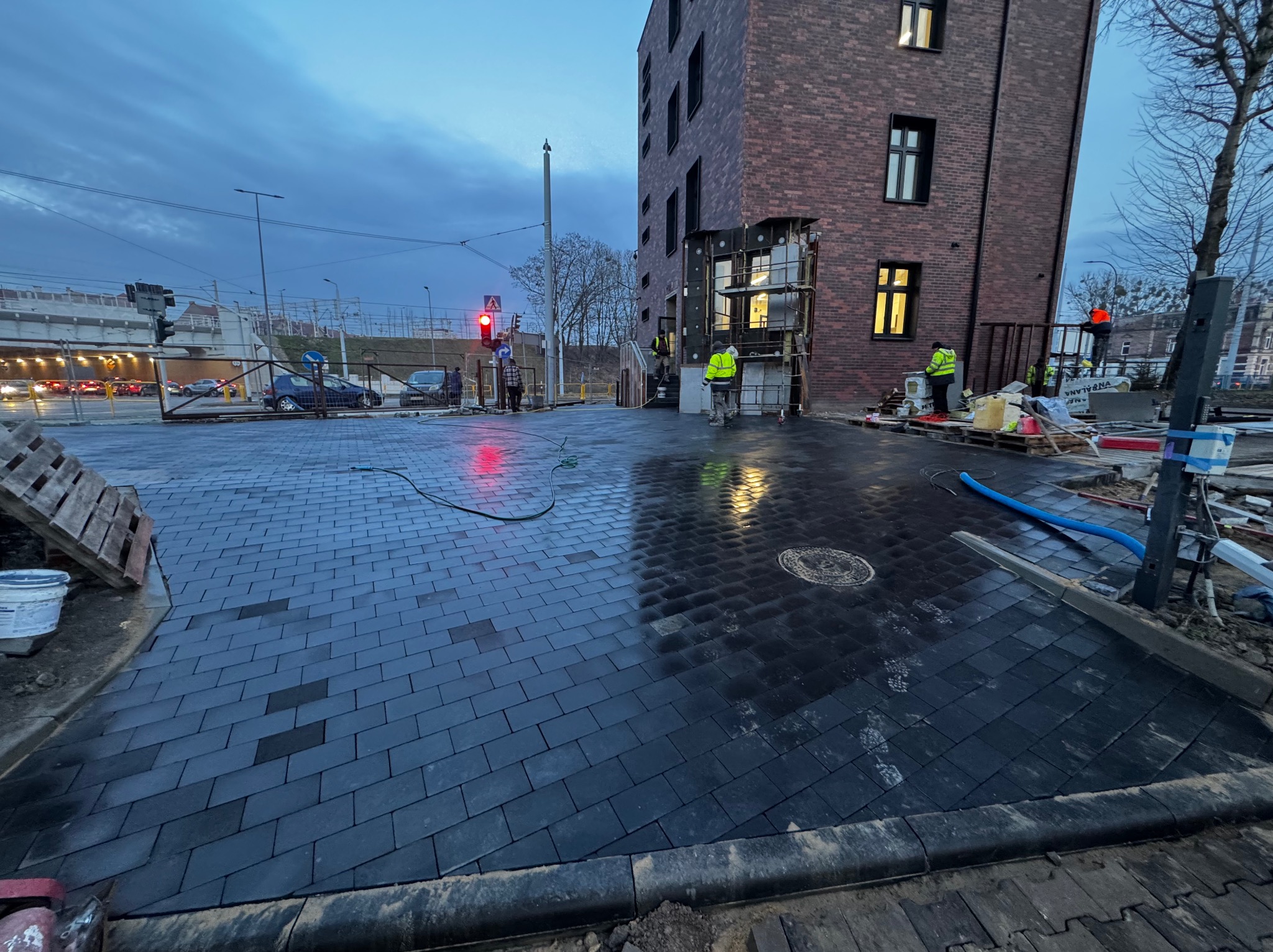 Ciemna kostka brukowa ułożona na placu budowy przy budynku z cegły. Widoczni pracownicy w odblaskowych kamizelkach i elementy infrastruktury miejskiej.