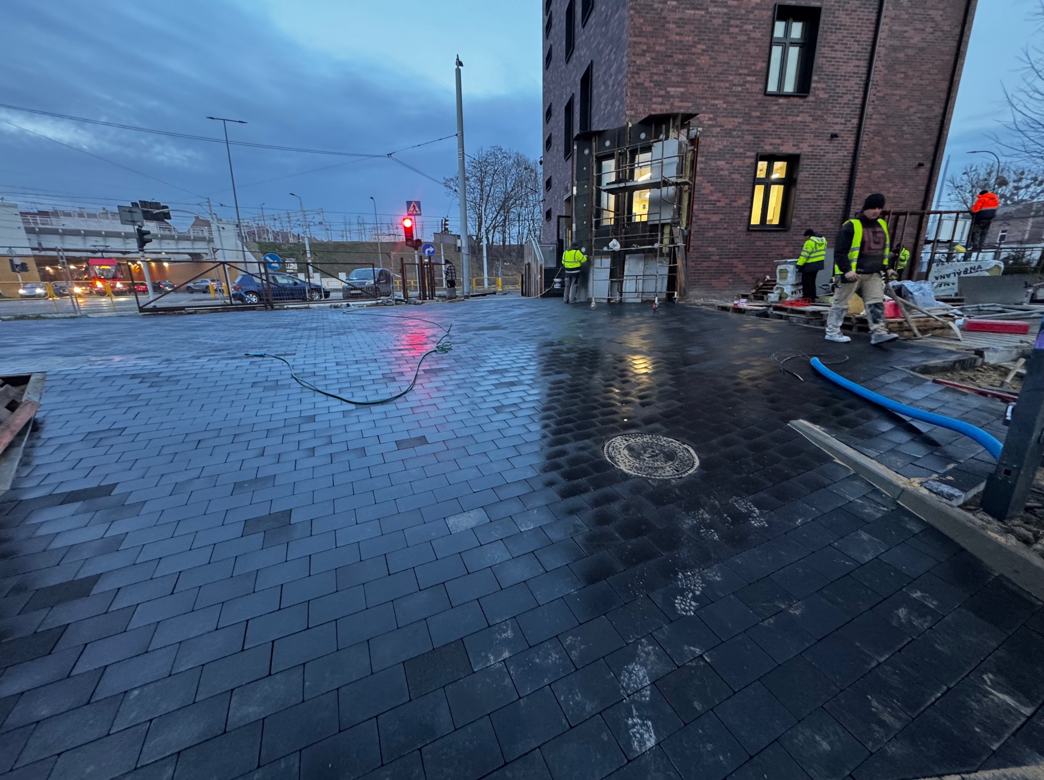Układanie ciemnej kostki brukowej na placu miejskim, widoczni pracownicy w odblaskowych kamizelkach, budynek w tle, sygnalizacja świetlna. Prace brukarskie w toku.