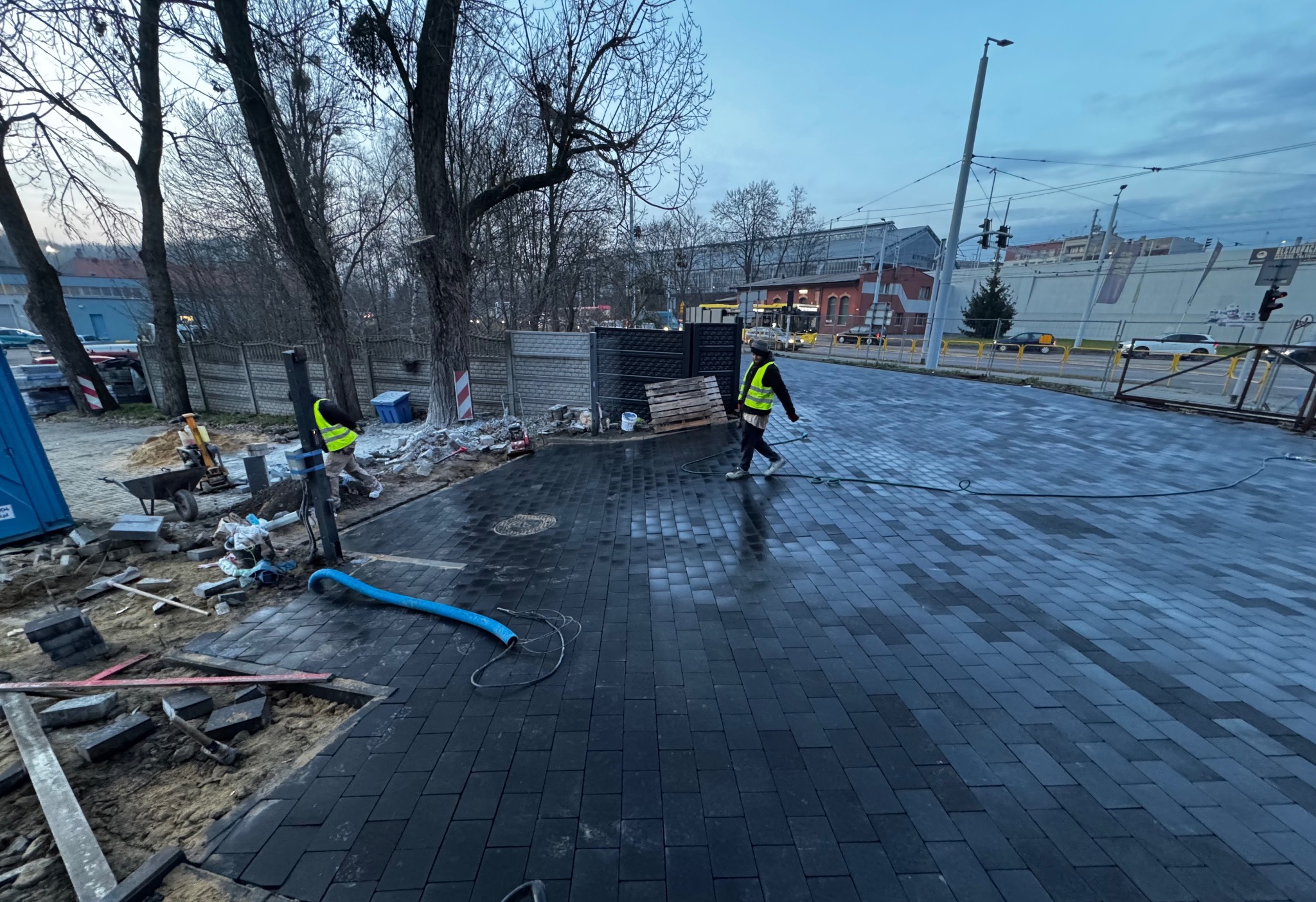 Ciemna kostka brukowa ułożona na chodniku miejskim, pracownicy w kamizelkach odblaskowych, widoczne narzędzia i materiały budowlane, tło miejskie z budynkami i drzewami.