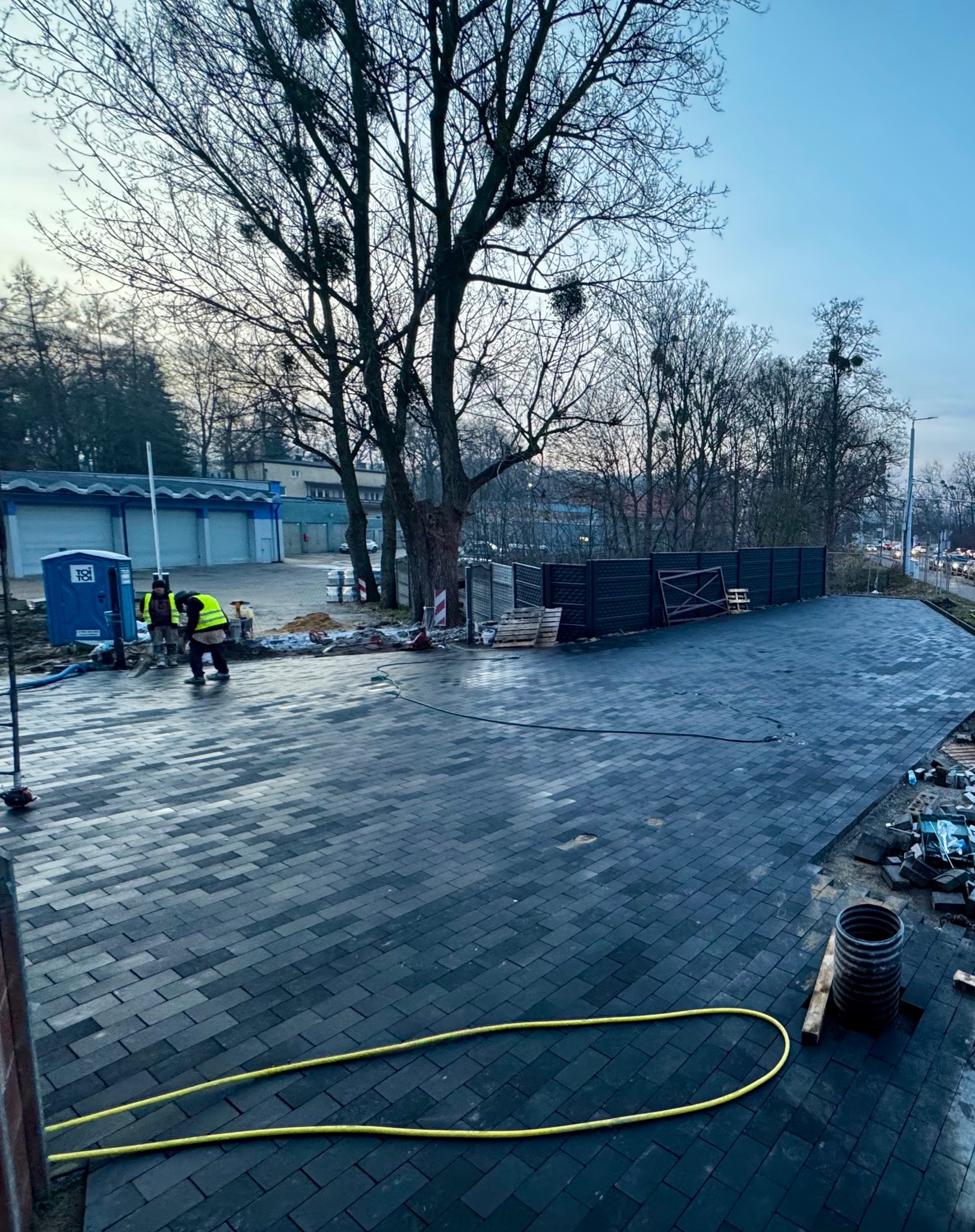 Ukończona nawierzchnia z ciemnej kostki brukowej na posesji, widoczni pracownicy w odblaskowych kamizelkach, fragment ogrodzenia i żółty wąż ogrodowy.
