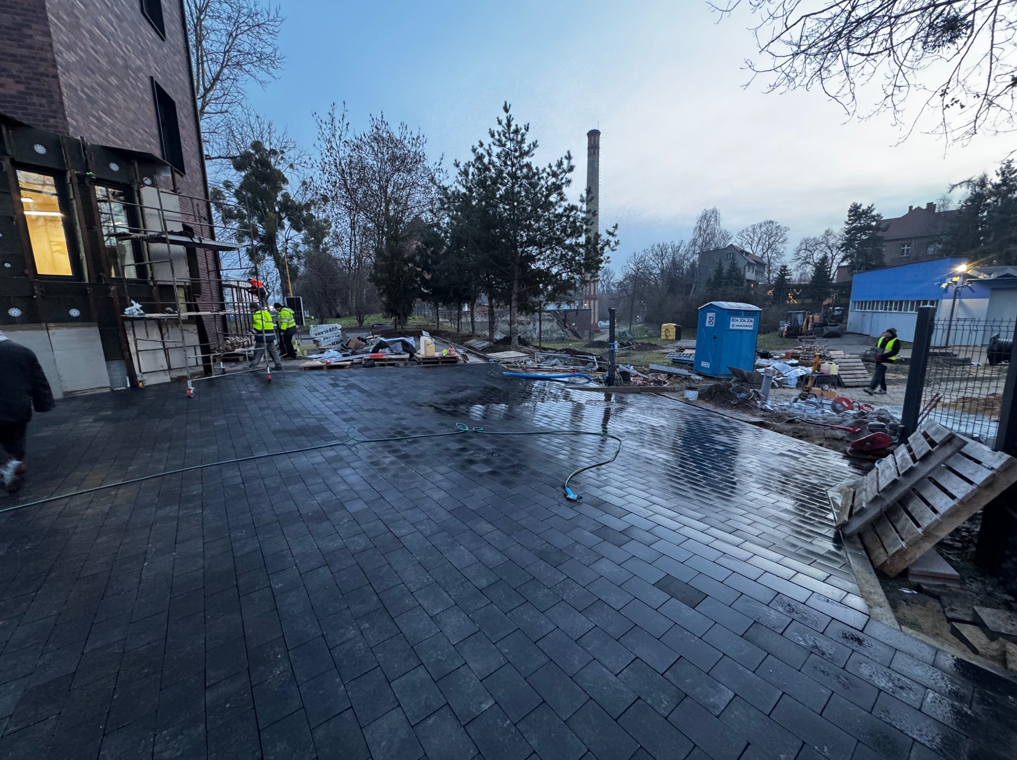 Ułożona, mokra, grafitowa kostka brukowa na posesji z budynkiem w tle. Widoczni pracownicy w odblaskowych kamizelkach i elementy placu budowy.