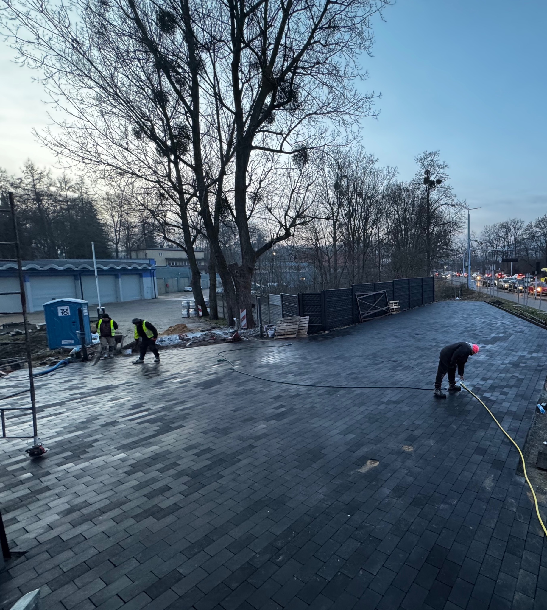 Ułożona, ciemnoszara kostka brukowa na placu, z pracownikami w odblaskowych kamizelkach i czerwonej czapce, myjącymi powierzchnię. W tle drzewa i budynki.
