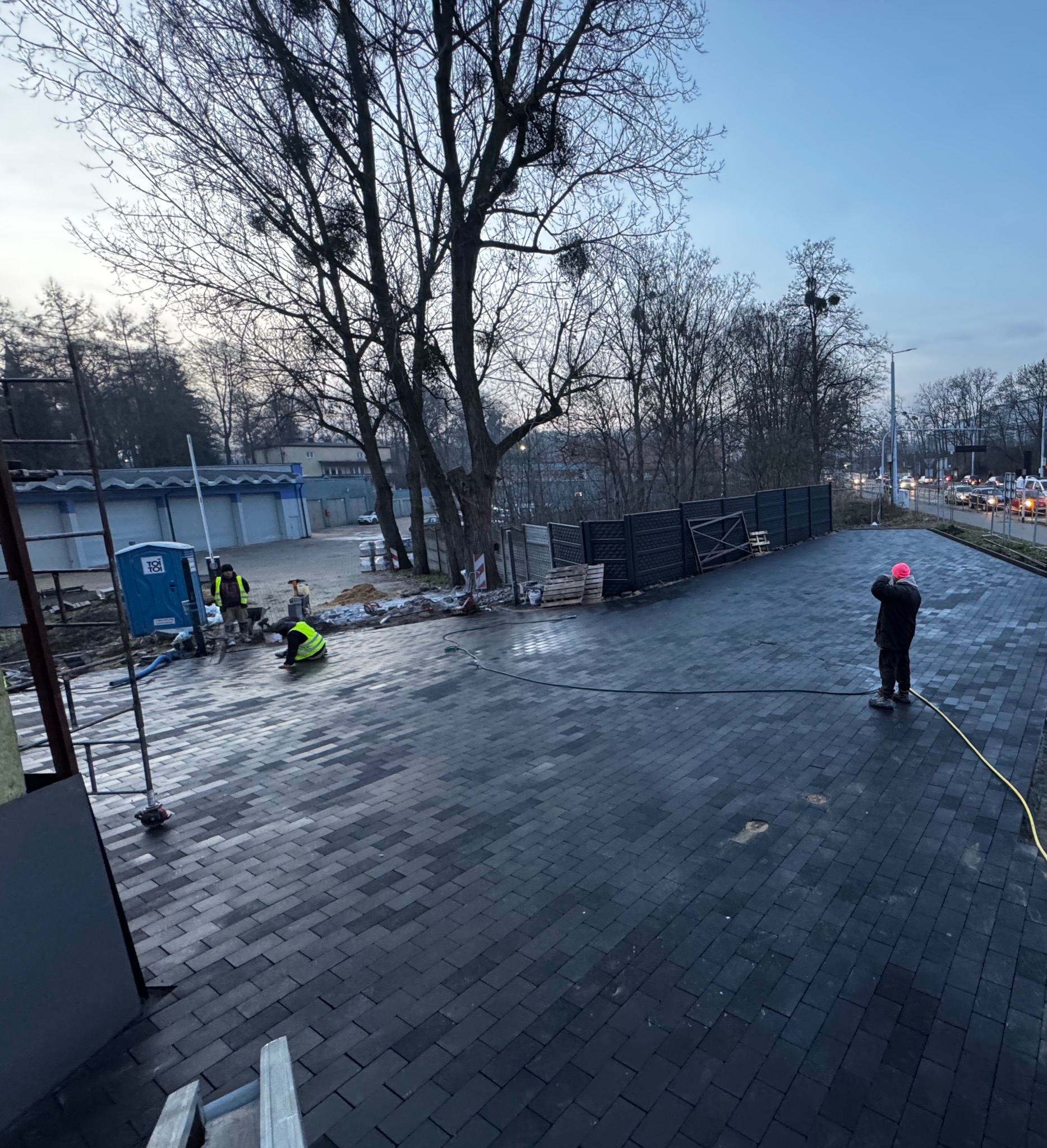 Ciemnoszara kostka brukowa ułożona na dużej powierzchni. W tle pracownicy w kamizelkach odblaskowych i budynek. Jeden z pracowników kontroluje jakość wykonania.