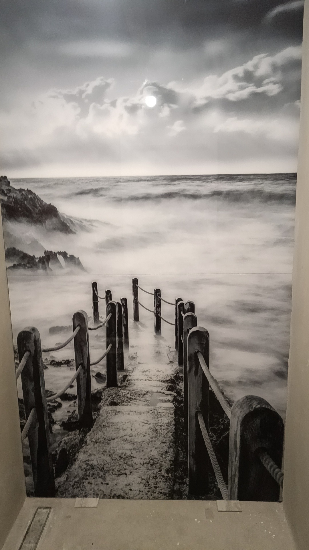 Czarno-biała fototapeta z motywem morskiego krajobrazu: pomost, fale i słońce na tle zachmurzonego nieba. Fototapeta naklejona na ścianie.