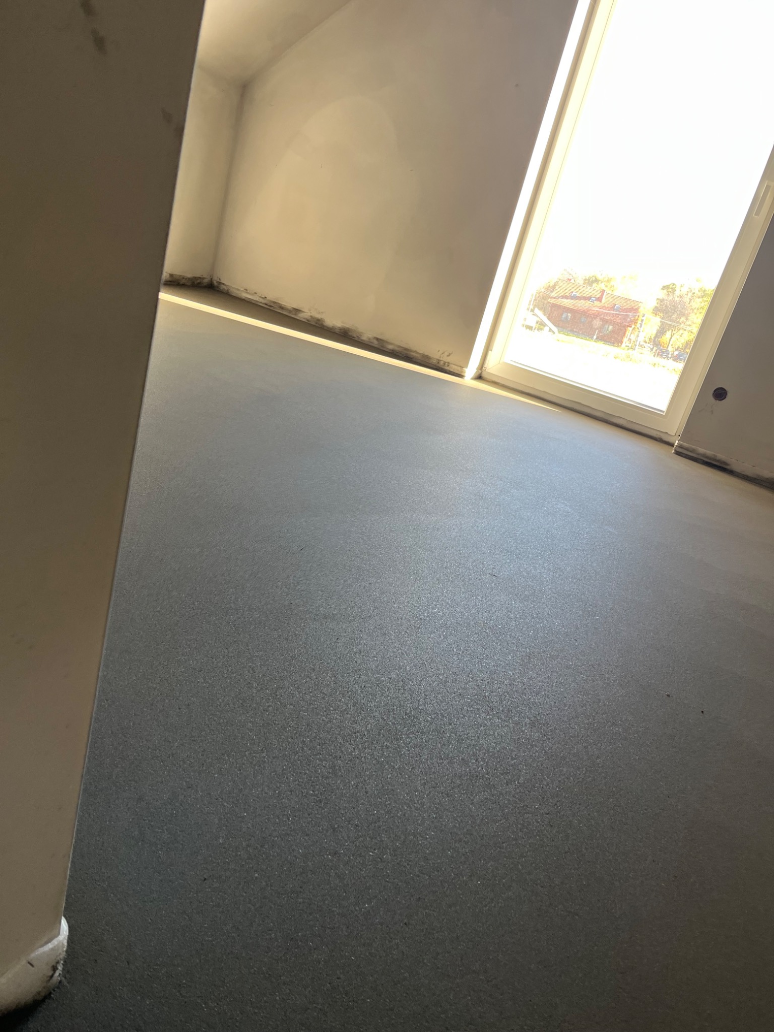 Szara, gładka posadzka w jasnym pomieszczeniu z dużym oknem. Widoczny fragment ściany i podłogi, z delikatnym światłem słonecznym wpadającym przez okno.