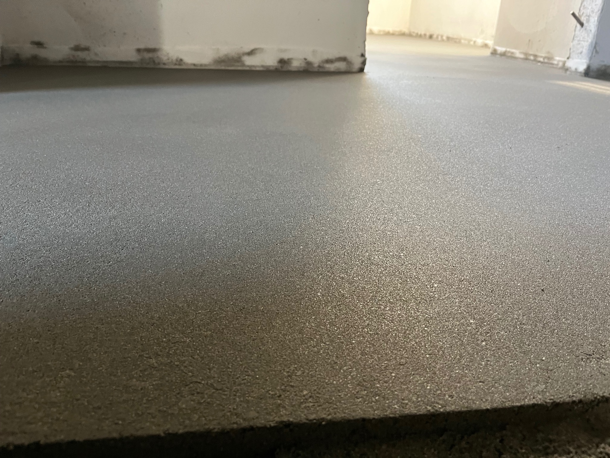Świeżo wylana, gładka posadzka w szarym odcieniu, z widocznymi śladami po narzędziach. W tle fragment ściany i rozproszone światło.