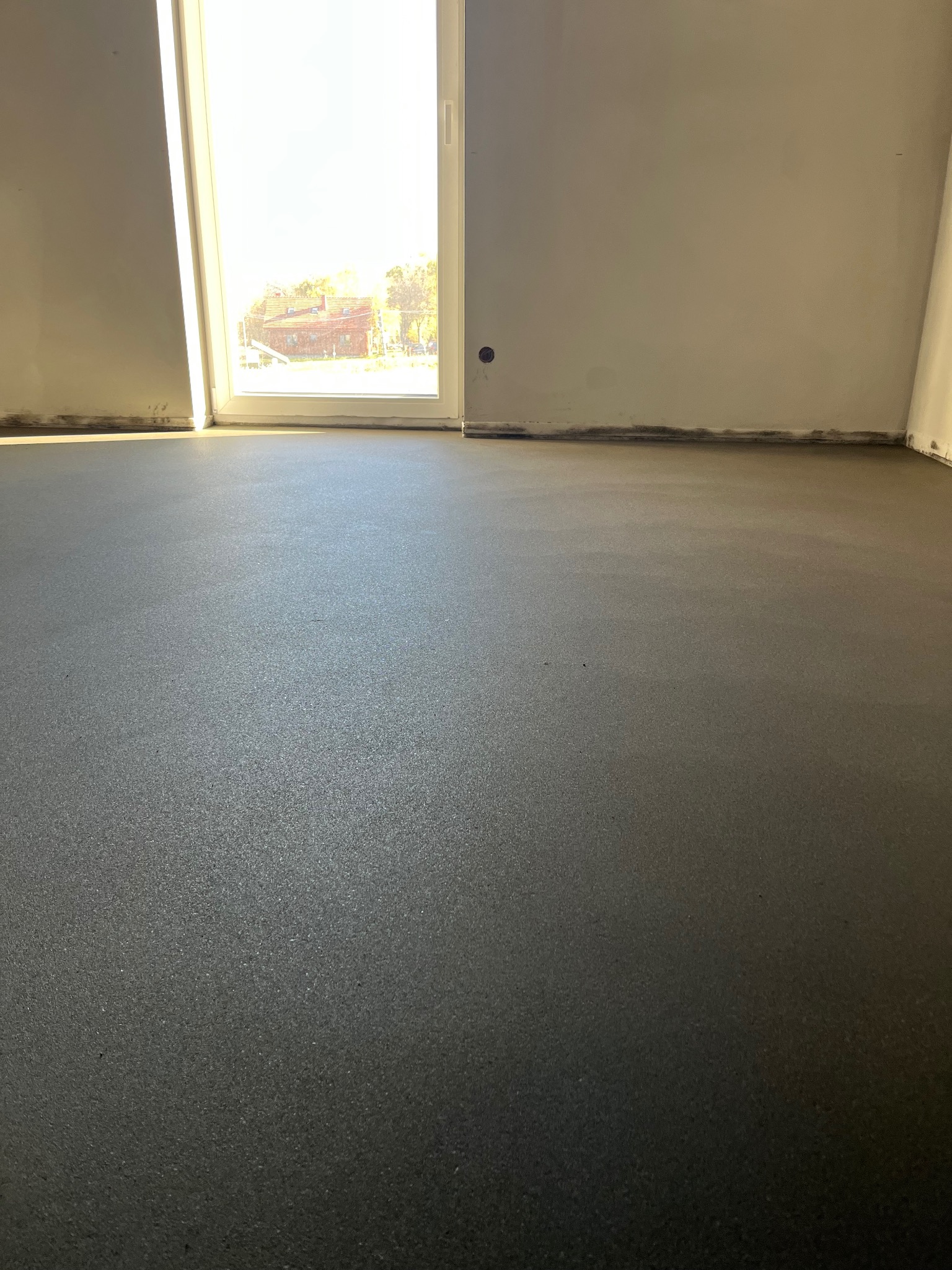 Świeżo wylana, gładka, ciemnoszara posadzka w jasnym pomieszczeniu z oknem. Widoczne ślady po poziomowaniu przy ścianach. Delikatne refleksy światła na powierzchni.