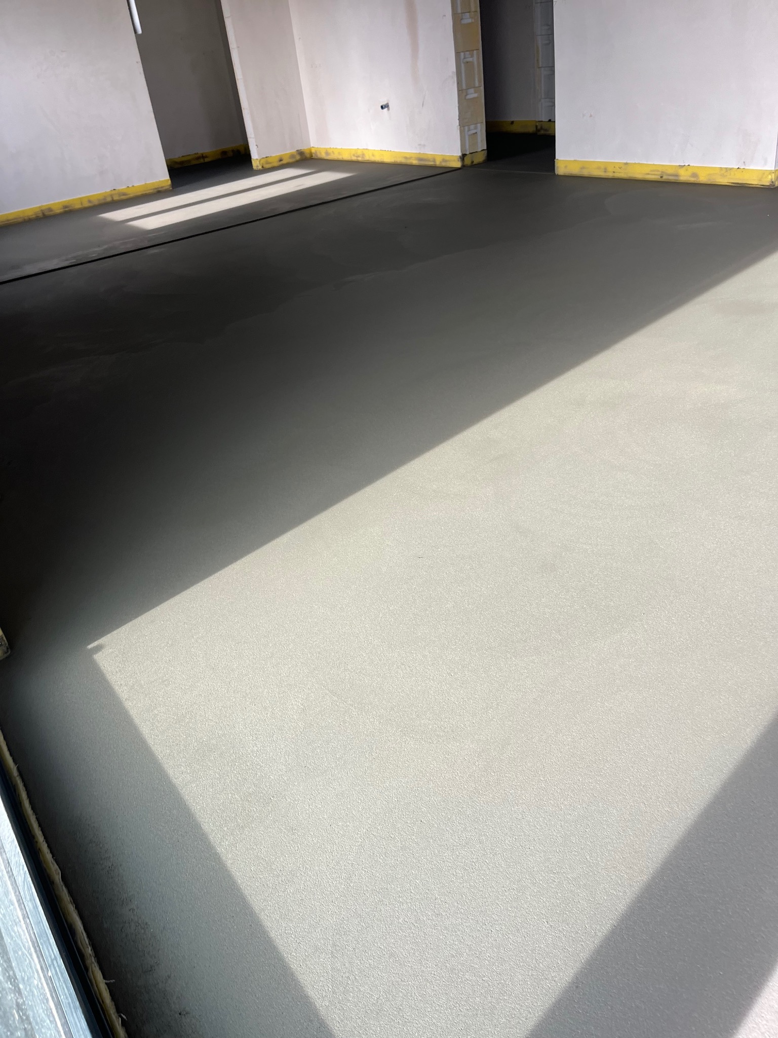 Świeżo wylana, gładka posadzka jastrychowa w jasnym pomieszczeniu z żółtymi paskami zabezpieczającymi przy ścianach. Widoczne ostre światło słoneczne.
